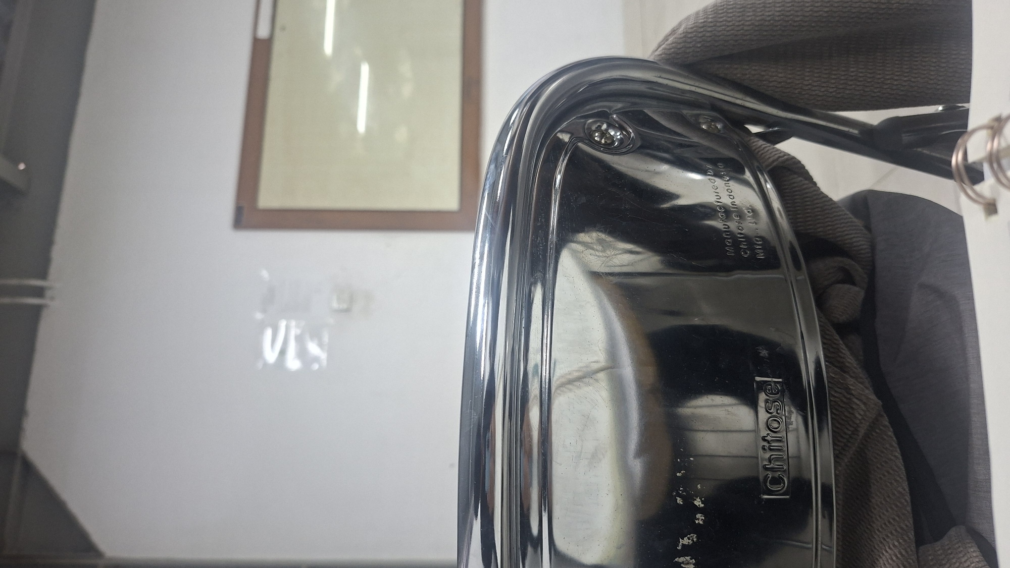 A close-up shot focuses on the shiny, curved chrome surface of what appears to be a chair's armrest or seat. Embedded in the chrome is a black plate with the embossed white text "Chitose" on the left. To the right of this, also in white embossed text, is "Manufactured by" followed by "Chitose Indonesia Mfg. Ltd.". A brown, textured fabric is draped over the lower portion of the object, obscuring the bottom. The background is out of focus, showing a white wall with a framed picture or bulletin board on the right, and the ceiling with fluorescent lighting fixtures above. The scene is indoors, likely in an office or public space in Padang, Indonesia, given the mention of "Chitose Indonesia Mfg. Ltd." on the object.