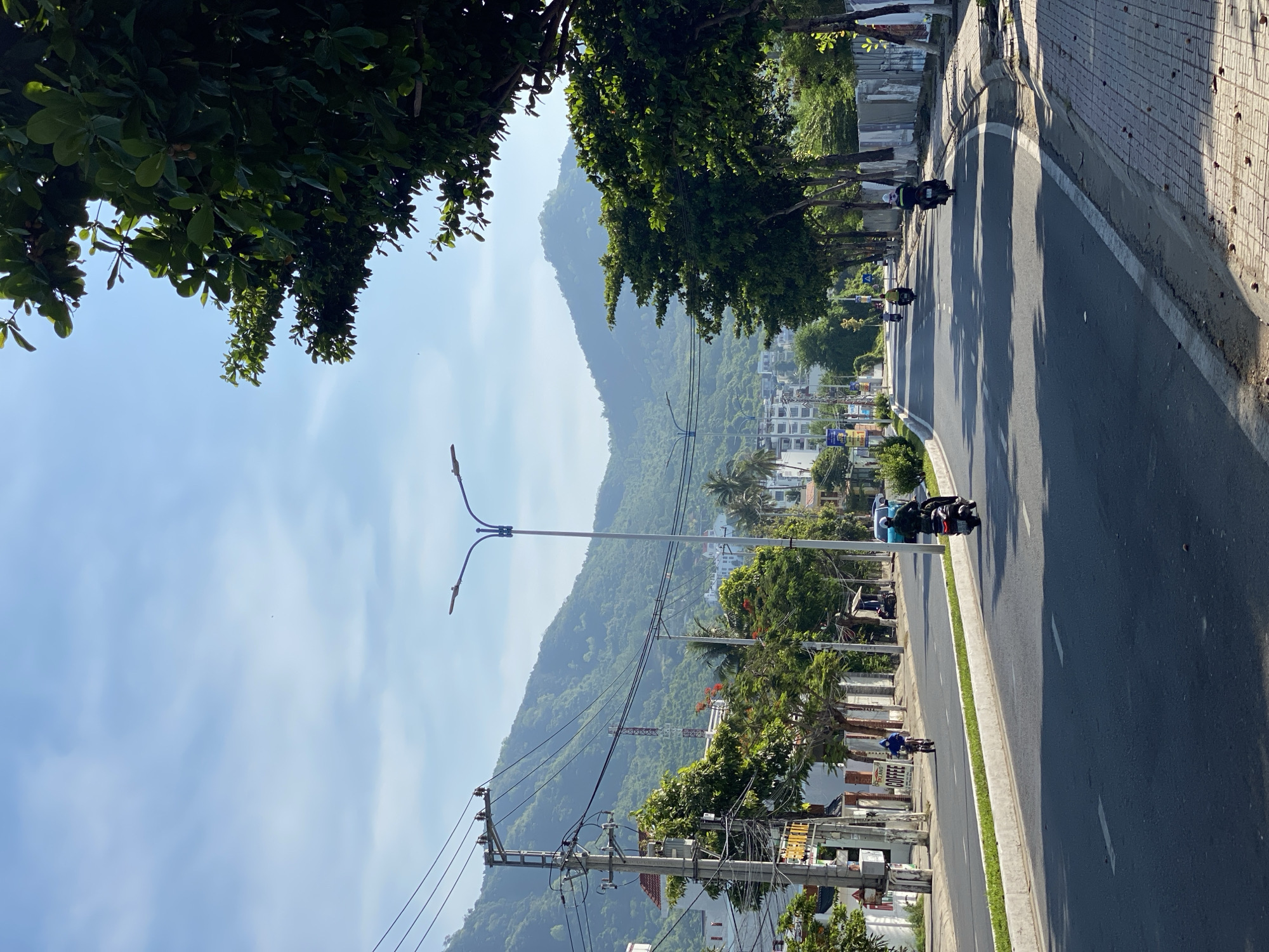 The image depicts a street scene in Bac Nha Trang Ward, Vietnam, on a clear, sunny day. A paved road runs through the center of the frame, with a green median strip separating lanes of traffic. On either side of the road are sidewalks, one paved with red bricks and the other lined with grass. Lush green trees, with dense foliage and some visible fruits, line the right side of the road, casting shadows on the pavement. In the background, a range of verdant, tree-covered mountains dominates the skyline. Buildings, some appearing to be apartment complexes or hotels, are visible nestled into the foothills of the mountains and along the left side of the road. Utility poles with numerous wires are present on the left side of the street, as are various signs, including one for "COFFEE." Several individuals are seen riding motorcycles on the road, with one person on a bicycle. The overall atmosphere is one of a bustling yet serene coastal town, bathed in bright sunlight.