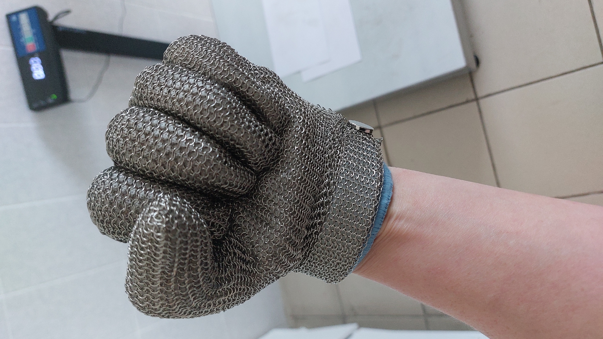 A close-up shot shows a person's forearm and clenched fist wearing a metal mesh glove. The glove is made of small, interlocking metal rings, forming a protective covering over the hand and fingers. The metallic texture is prominent, with visible individual links. The wrist of the glove has a blue fabric cuff. The forearm is bare and shows skin tone. In the background, a digital scale with a display showing "003" is visible, along with a tiled floor and a portion of a white surface. The lighting is bright, casting subtle shadows. There is no indication of the city of Yelizovo, Russia, in the image.