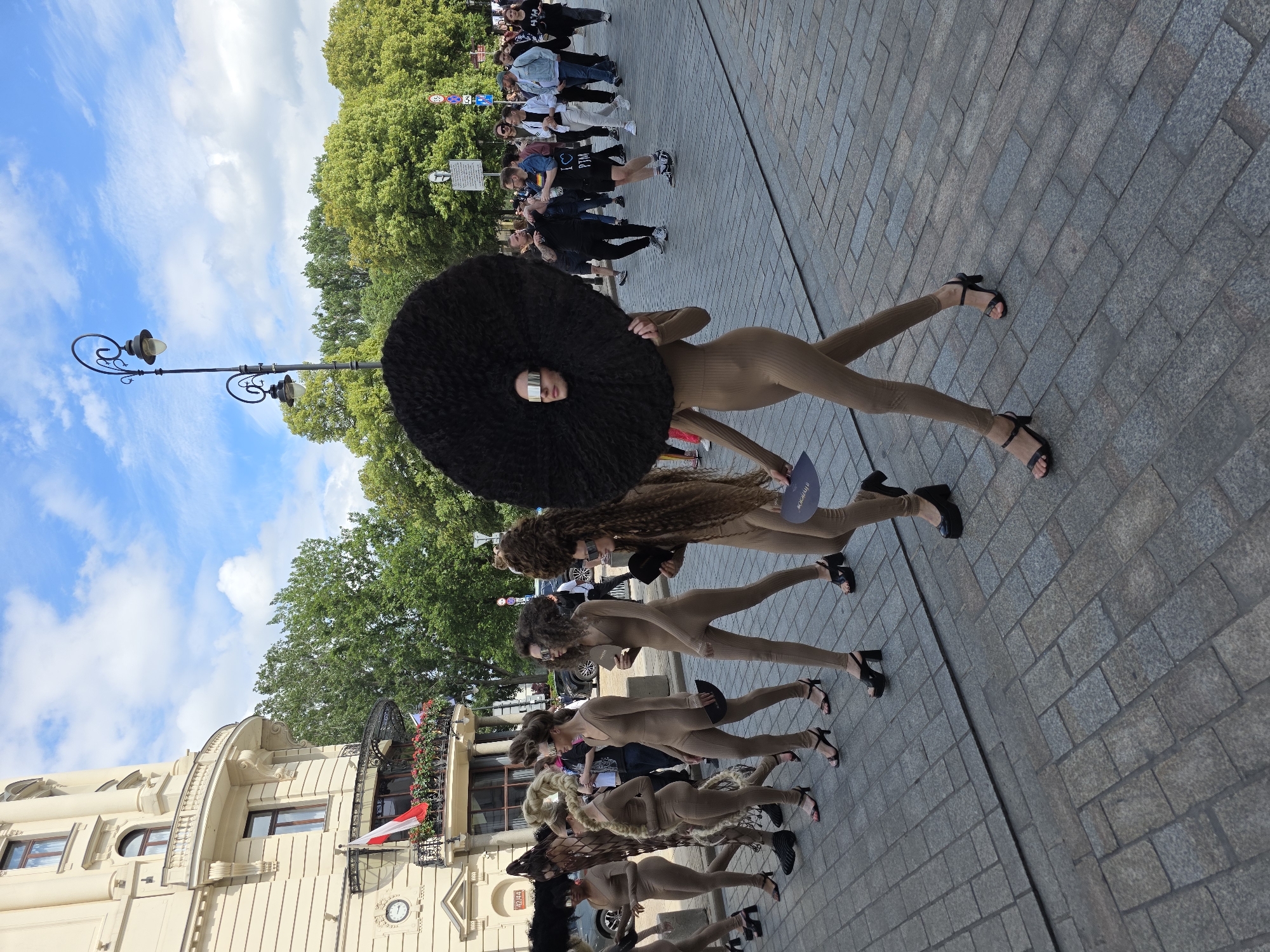 A group of people are walking in a parade down a cobblestone street in Warsaw, Poland. The people in the foreground are dressed in tight-fitting beige outfits and elaborate, curly wigs. The person in the very front has a large, dark, circular afro wig and wears silver mirrored glasses. The people behind them have various styles of wigs, some long and blonde, others shorter and brown, all with a similar curly texture. They are all wearing high-heeled sandals. The background shows a building with classical architecture, trees, a street lamp, and other onlookers. The sky is blue with scattered white clouds. The scene appears to be a fashion show or cultural event.