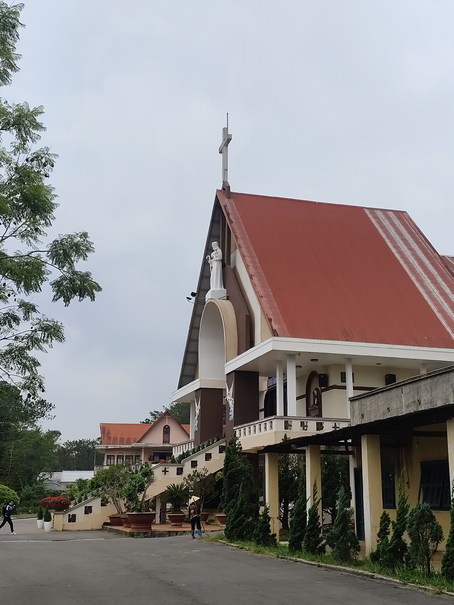 The image depicts a Catholic church in Lang Biang - Đà Lạt, Vietnam. The church features a prominent red metal roof with a cross atop the gable. A white statue stands above a large arched entrance. Adjacent to the church, a lower building with yellow walls and a covered walkway is visible. The foreground shows a paved area with small coniferous trees lining the edge. In the background, a smaller building with a similar red roof can be seen. Two people are present in the image. One is walking along the paved area on the left, and the other is sweeping or raking near the stairs leading up to the church. The sky is overcast. Potted plants are placed along the stairs leading to the church.