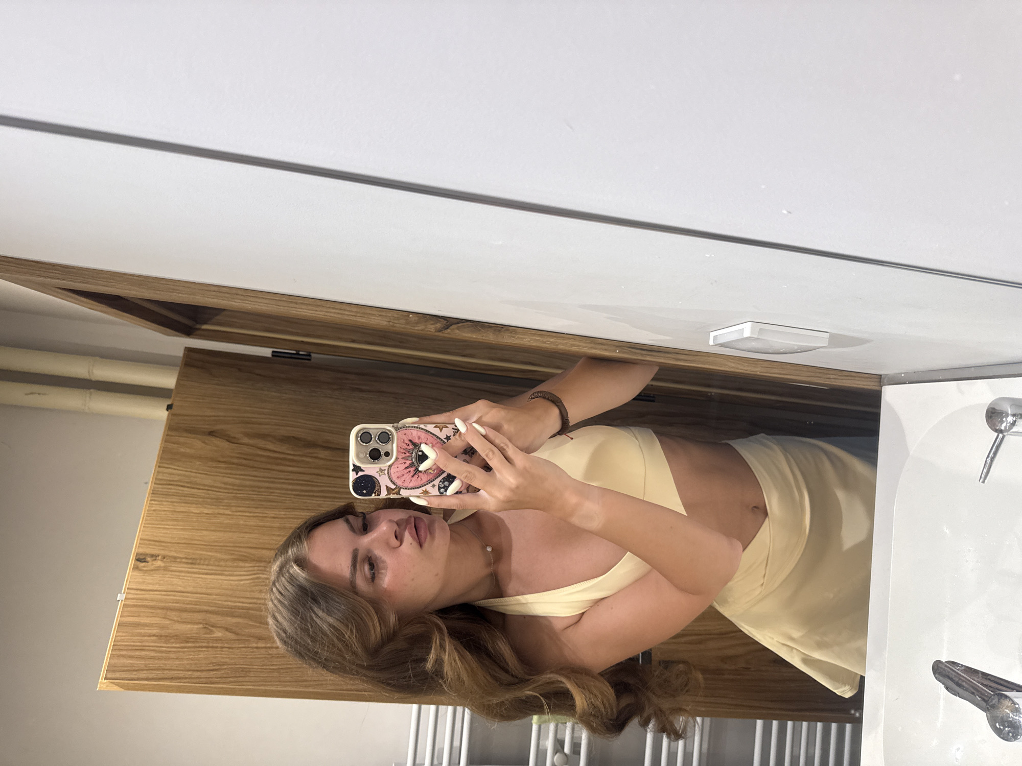 In a bathroom, a young woman with long, wavy brown hair takes a selfie in a mirror. She is wearing a light yellow halter-neck crop top and a matching skirt. Her phone, with a pink case adorned with stars and a heart, is held up to capture the reflection. The background reveals a wooden door, a white wall, and a sink with faucets. This scene is located in the city of Plovdiv, Bulgaria.