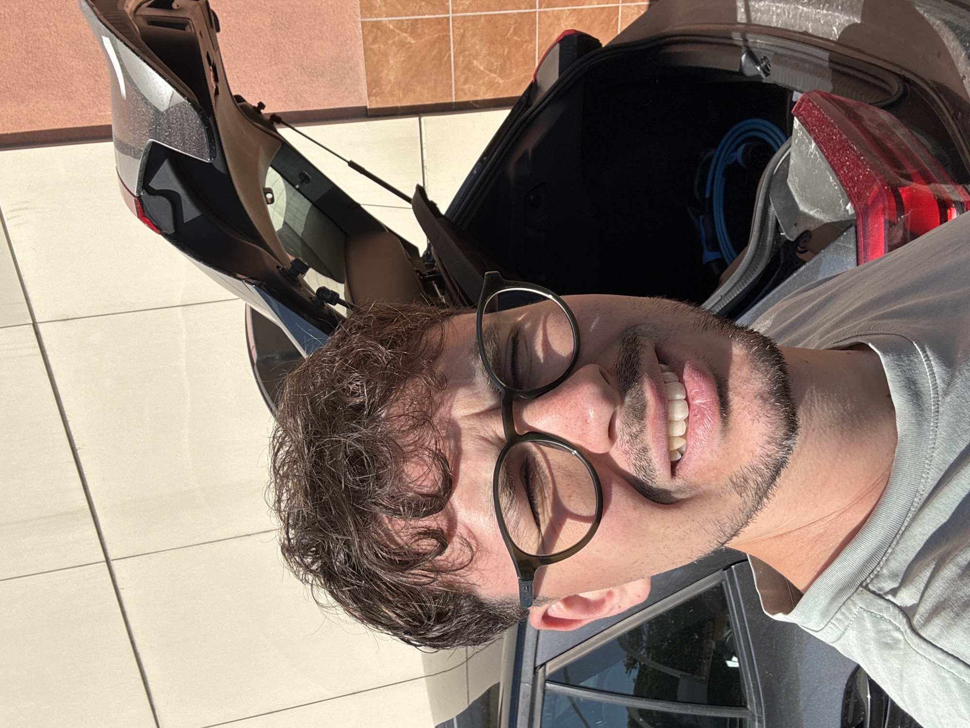 A man wearing round glasses and a grey shirt is standing next to an open car trunk. The car is parked on a tiled surface. The background shows some buildings, consistent with an urban environment. There is a blue cord inside the trunk. The image is oriented vertically. Based on the tiles and architecture, the location is consistent with Varna, Bulgaria, but without further evidence this cannot be determined with certainty.
