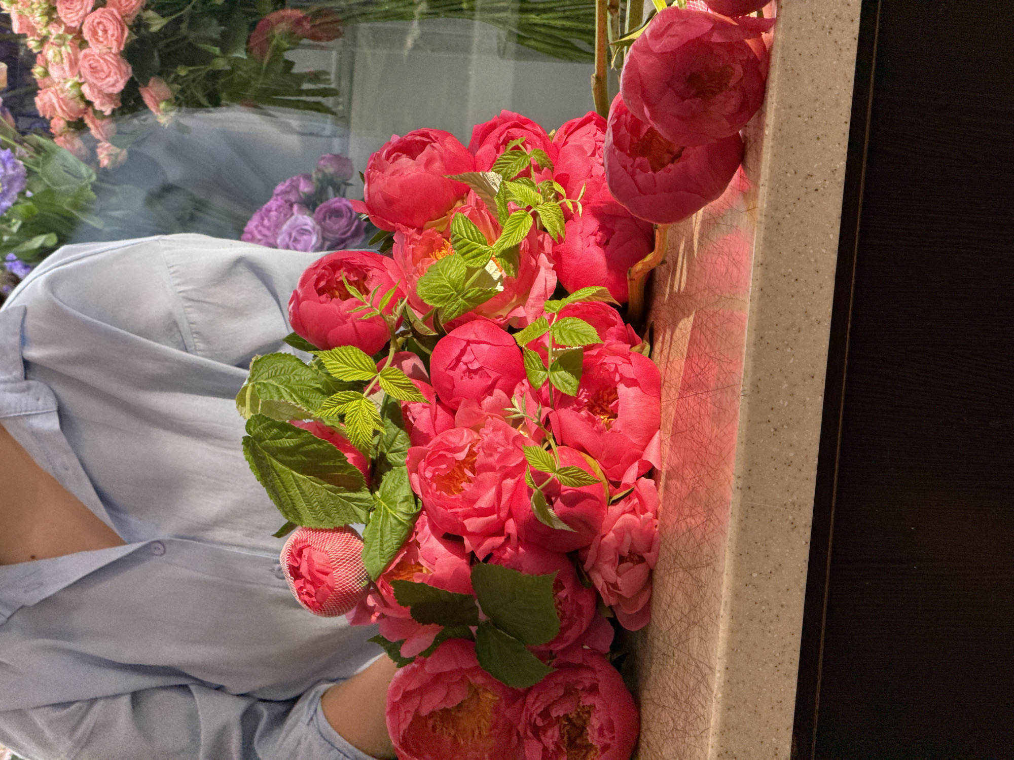 A person in a light blue collared shirt holds a large bouquet of vibrant pink peonies. The peonies are in various stages of bloom, with some fully open and others still tightly budded. Green leaves are interspersed throughout the bouquet. The bouquet rests on a speckled beige countertop, which has a series of faint scratches. Behind the bouquet, a blurred reflection shows other flowers, including pink roses and purple flowers, suggesting a flower shop or floral arrangement setting. The background is out of focus but hints at glass surfaces and more floral displays. The scene is illuminated by natural light, casting shadows and highlighting the rich colors of the flowers. There is no information provided about the location being Assinovskaya, Russia.