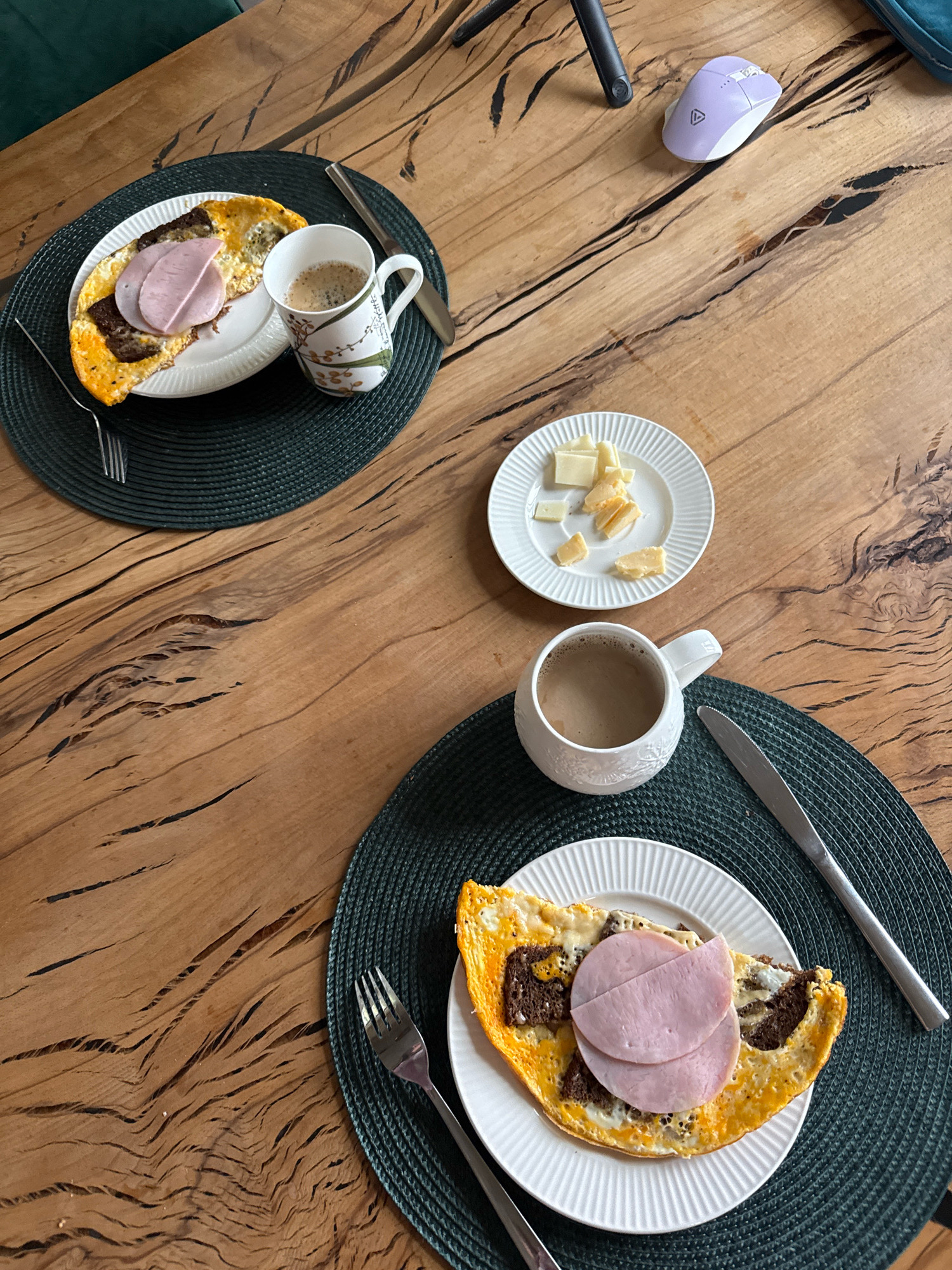 The image shows a wooden table with two place settings for breakfast. Each place setting includes a white plate with an omelet topped with ham and pieces of dark bread, a white mug of coffee, a dark green placemat, and silverware. There is also a small white plate with pieces of cheese on the table. In the upper right corner of the image, a light purple computer mouse is visible on the wooden table. The table surface has a distinctive grain pattern with visible cracks and lines. The overall scene depicts a breakfast meal laid out on a rustic wooden table. The location is not specified in the image.