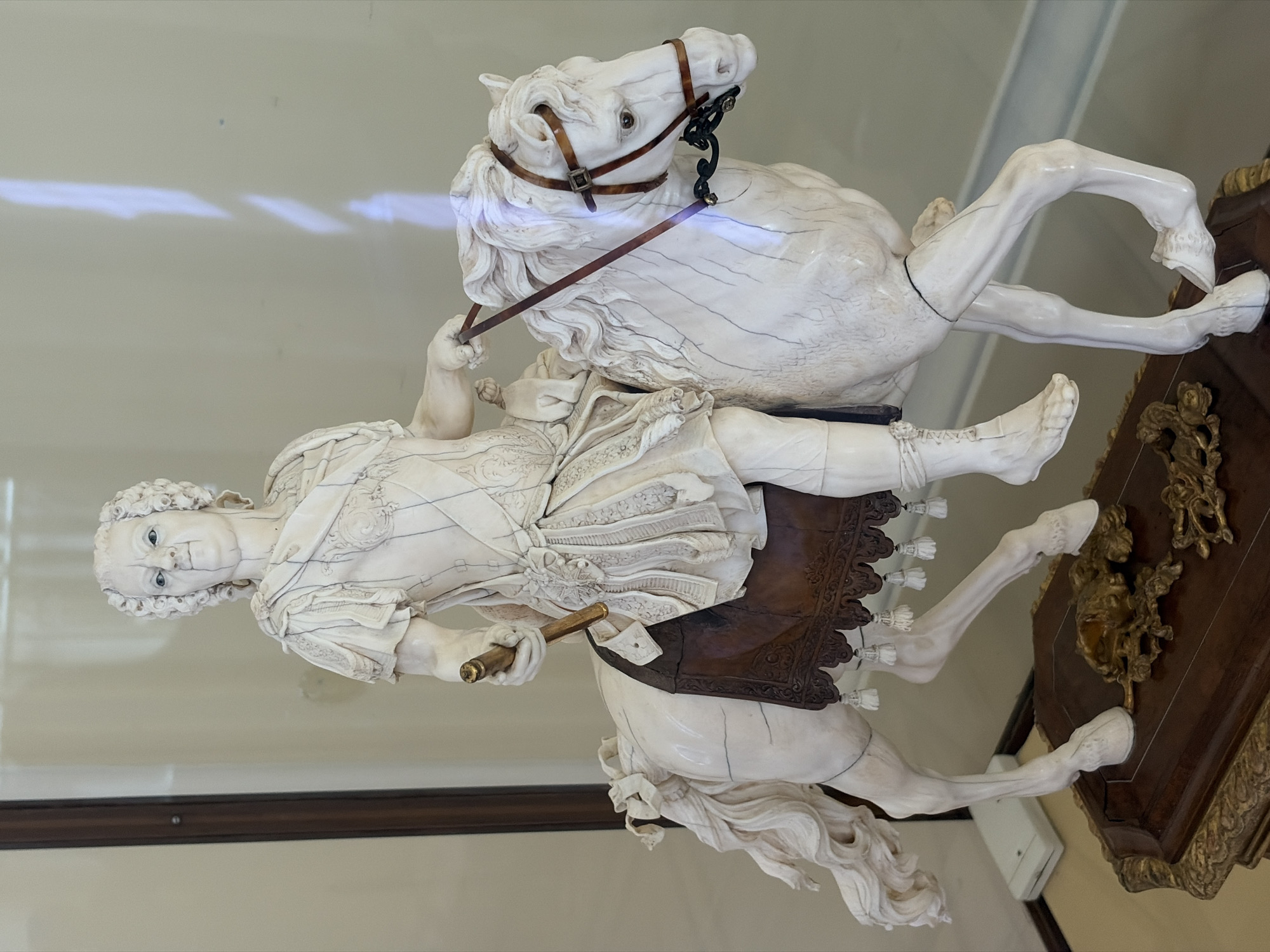 The image depicts an antique ivory sculpture of a man on horseback displayed in a glass case. The sculpture is intricately detailed, with the man dressed in period clothing and the horse equipped with a bridle and saddle. The sculpture's patina suggests age. It is located in Saint Petersburg, Russia, within the Hermitage Museum, identified by similar items housed there.
