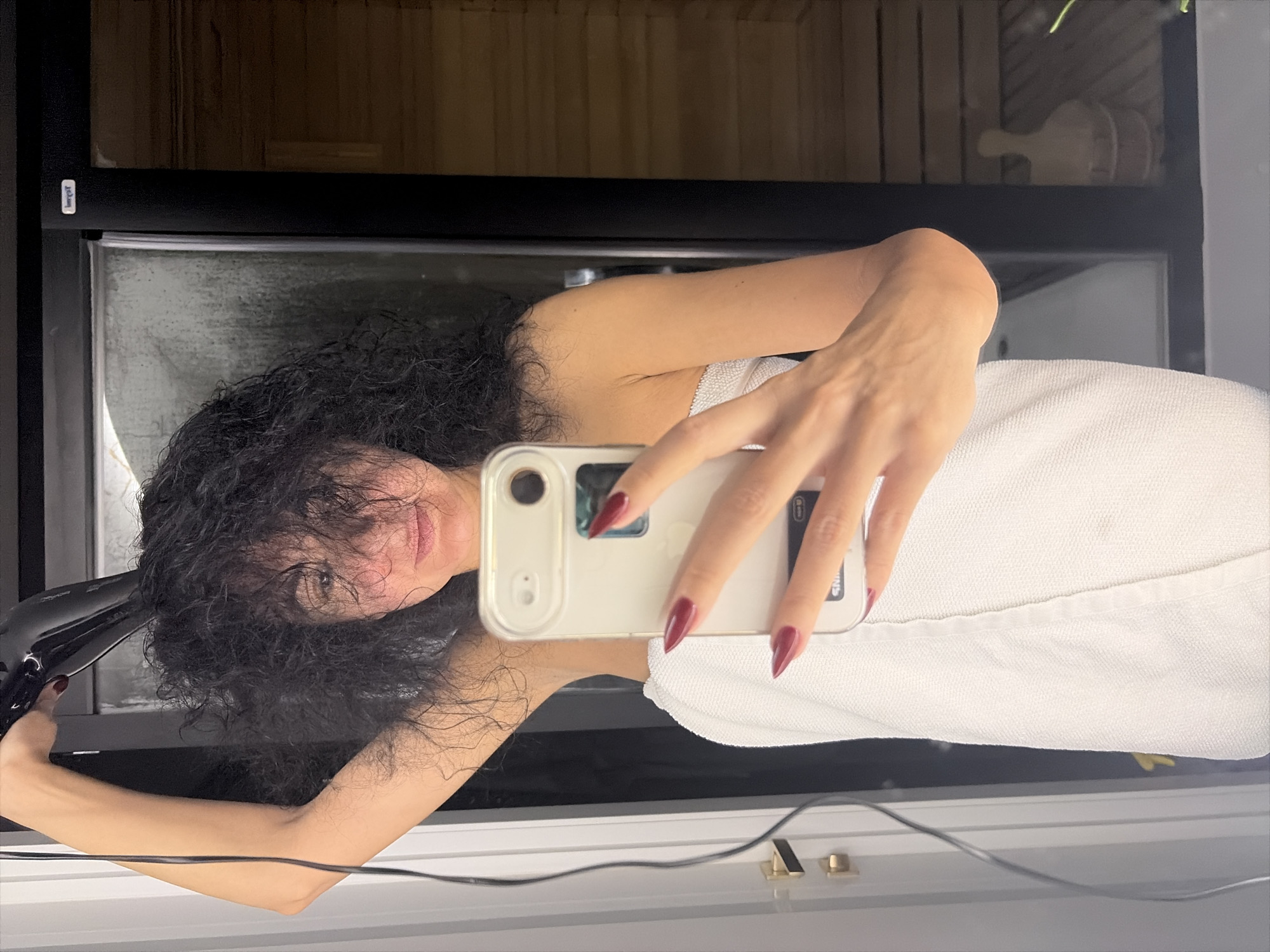 The image shows a woman in a bathroom, possibly in Moscow, Russia, drying her long, dark, curly hair with a black hairdryer. She is wrapped in a white towel and holding a silver smartphone in her right hand to take a selfie in a mirror. The background reveals a glass door with wooden paneling, possibly indicating a sauna or steam room.