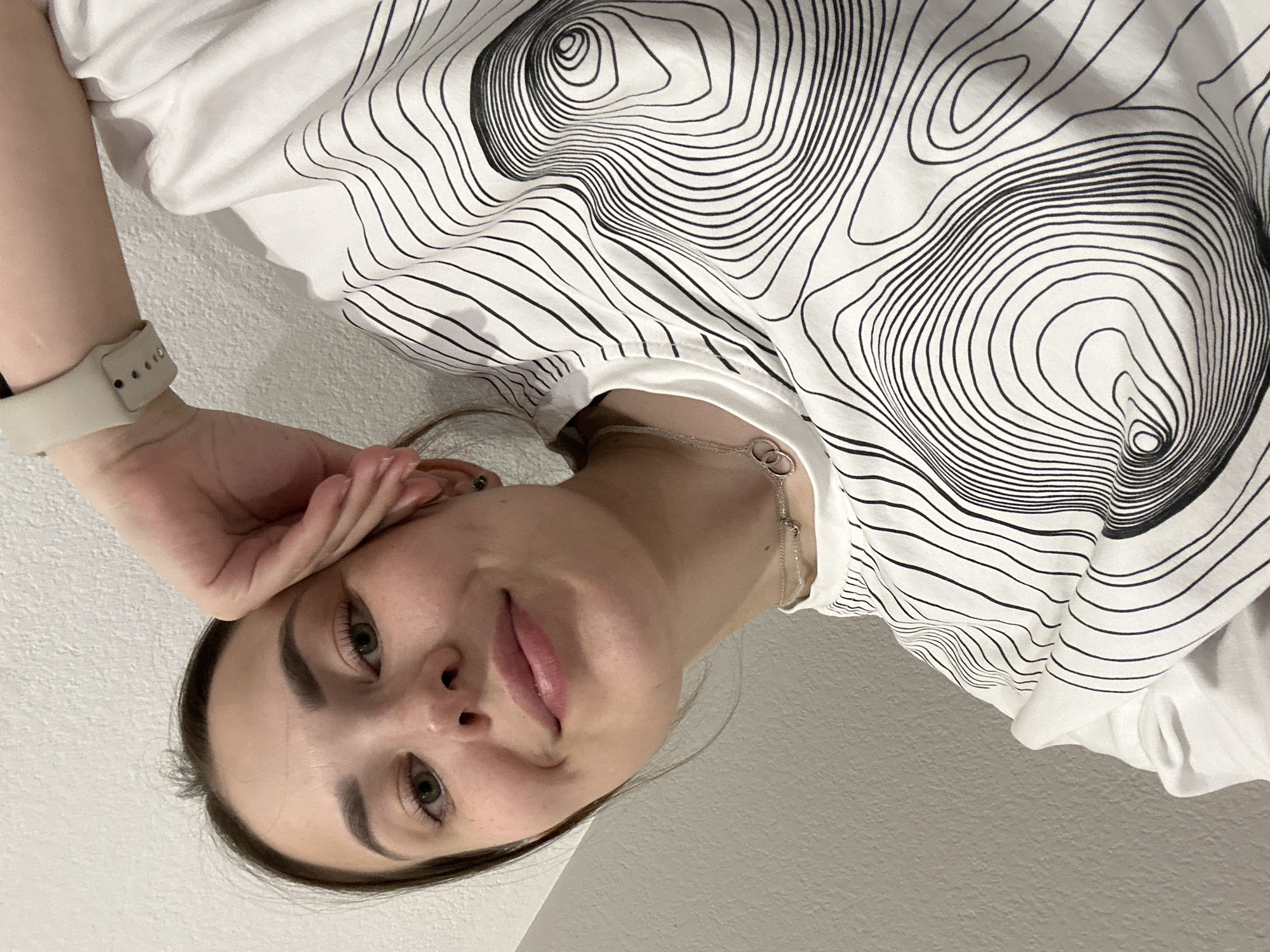 A woman poses for a selfie. She is fair-skinned with green eyes, brown hair, and defined eyebrows. She is wearing a white T-shirt with a black topographic-style pattern. A silver chain necklace is visible around her neck. She has a light-colored smartwatch on her left wrist. Her left hand is gently touching her face near her ear. The background is a plain white wall. There is no evidence in the image to suggest it was taken in Perm, Russia.