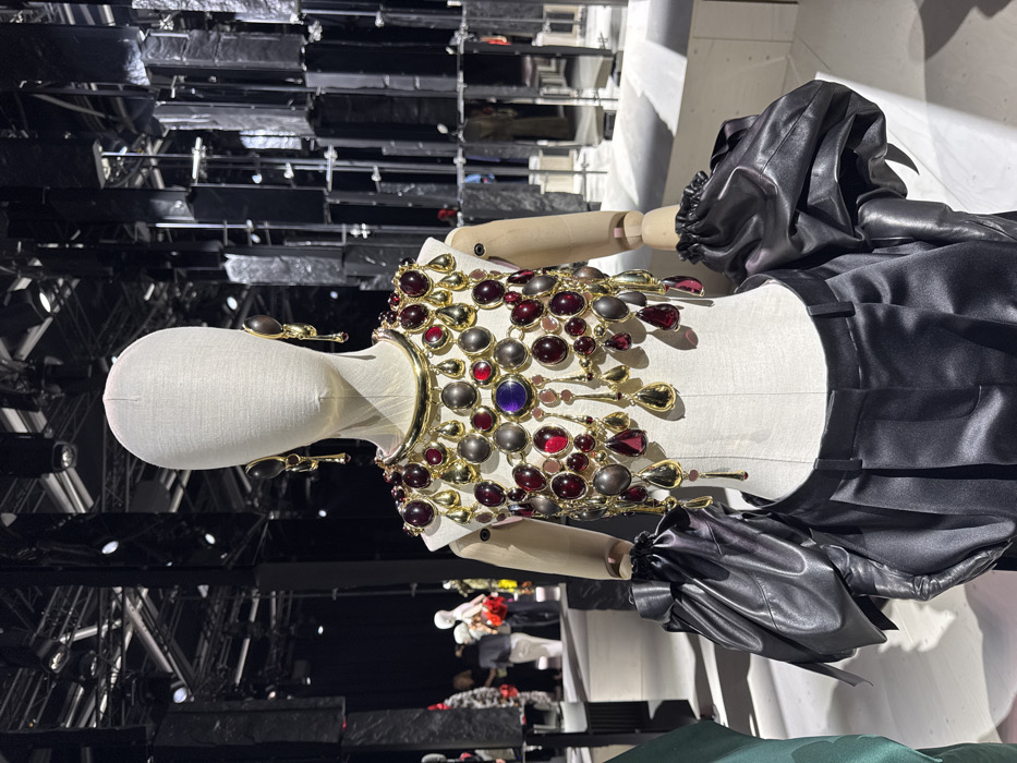 A headless, fabric-covered mannequin with articulated wooden arms stands center, displaying a high-fashion ensemble within a museum or exhibition space. The mannequin wears an ornate, gold-toned metal crop top heavily embellished with numerous cabochon-cut red and dark gray stones, with a single purple stone at the center. Gold-toned teardrop pendants hang from the top. A rigid, smooth gold-toned choker encircles its neck, and large, elongated gold-toned dangle earrings with dark, oval elements adorn its ears. Voluminous black, shiny, possibly faux-leather or satin puff sleeves, appearing to integrate with long black gloves, cover its arms. High-waisted, pleated black satin trousers complete the outfit.
The environment is characterized by a dark ceiling with visible spotlights and structural elements. In the blurred background to the left, indistinct figures of visitors and other display mannequins are visible. To the right, a striking architectural installation features multiple vertical, rectangular, fragmented reflective panels in a dark material, creating a complex visual effect. The floor is a light, possibly white or pale gray, polished surface. This scene is located at the Musée des Arts Décoratifs (MAD Paris), 107 Rue de Rivoli, Paris, France.