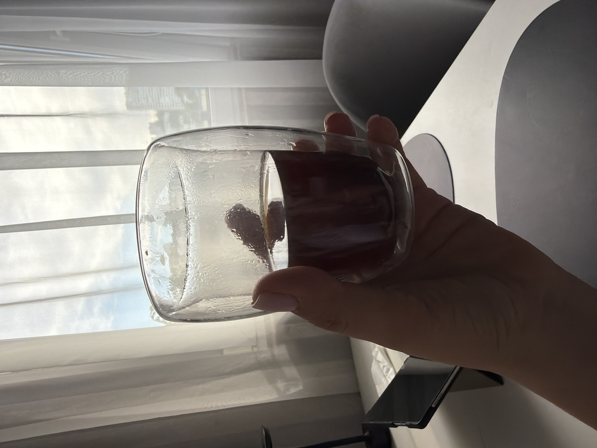 A first-person, eye-level indoor shot depicts a hand holding a double-walled glass against a brightly lit window overlooking an urban landscape in the city of Sochi, Russia.
In the foreground, a human hand with light pink-polished fingernails grasps a clear, double-walled glass. The glass exhibits condensation on its exterior, indicating a temperature differential. Inside, a dark, reddish-brown liquid, consistent with a beverage, contains at least one whole raspberry and a thin, yellowish slice of citrus fruit, possibly lemon.
The hand and glass are positioned over a white or light-colored table. On the table's left side, a dark-colored tablet device rests in an upright stand. To the right of the hand, a dark gray, semi-circular object, resembling a placemat or coaster, is visible on the table. A similar, partially obscured circular object is positioned beneath the glass. Further to the right, a dark gray, rounded chair back is partially visible.
The background is dominated by a large window covered with sheer, light-colored curtains that are drawn to the sides, allowing significant natural light to enter. Through the window, a bright, overcast sky is visible, along with several multi-story urban buildings in the distance, partially obscured by the curtains and atmospheric conditions. A dark, vertical element, possibly a lamp stand, is present on the far left edge of the frame. The overall lighting is natural and bright, silhouetting the foreground elements against the luminous exterior view.
