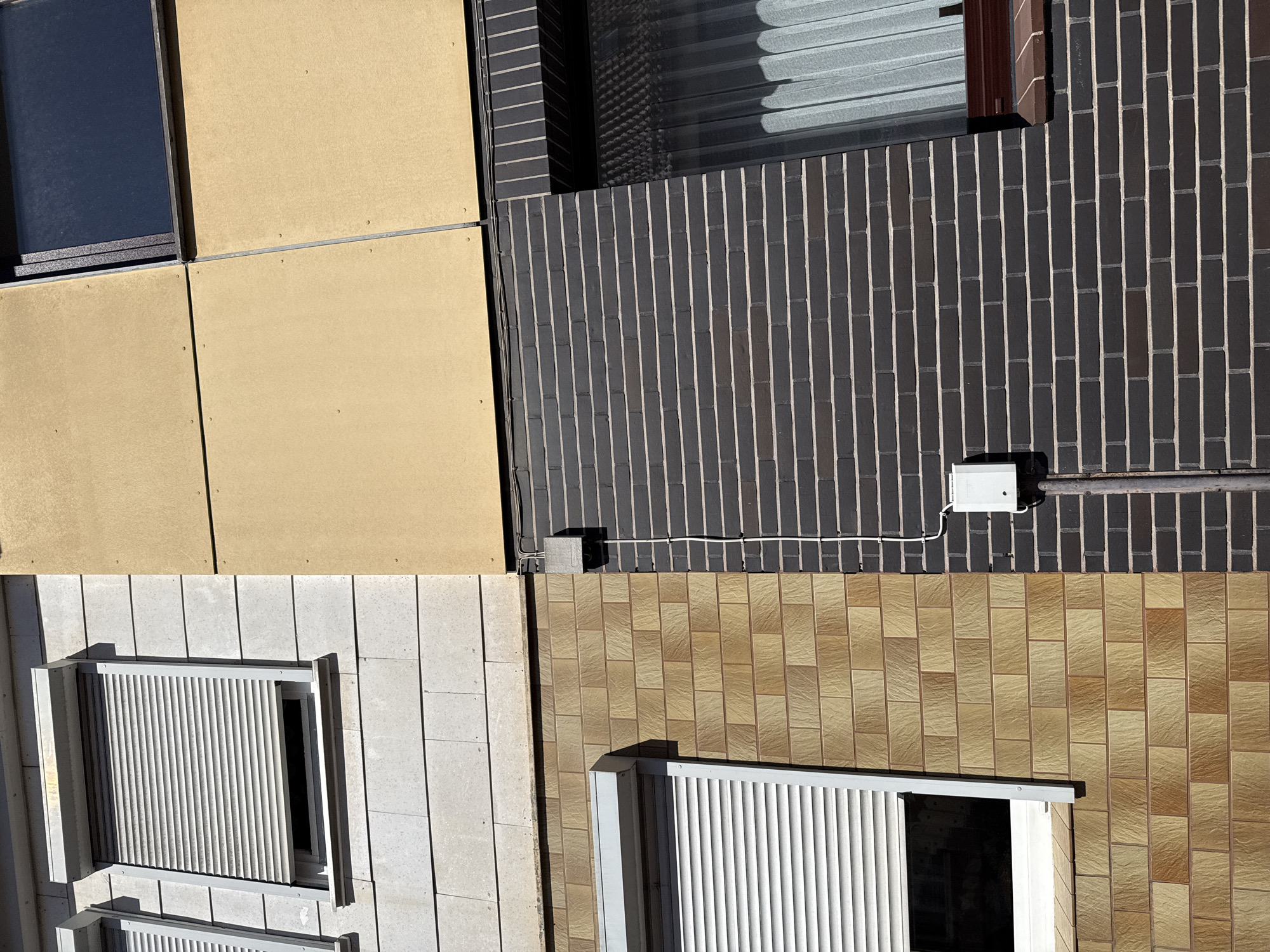 The image shows the exterior facade of a building located in the city of Bad Sachsa, Germany. The facade is divided into sections with different materials and colors. On the left, a section of the building features a window with a gray roller shutter, set against a light gray, textured stone or tile cladding. To the right of this, a large section of the building is covered in a matte yellow, paneled material. Below the yellow panels and to the right of the light gray cladding, there is a window with a closed roller shutter, framed by dark brown brickwork. Further to the right, a dark gray brick wall dominates the lower portion of the image, with a window covered by sheer white curtains visible on the far right. An electrical junction box is mounted on the dark gray brick wall, with white cables running down to a conduit pipe. The building appears to be a multi-story structure, possibly residential or commercial, with clear delineation between the different construction materials. The lighting suggests it is daytime, with sunlight casting shadows. There are no people or animals visible in the image.