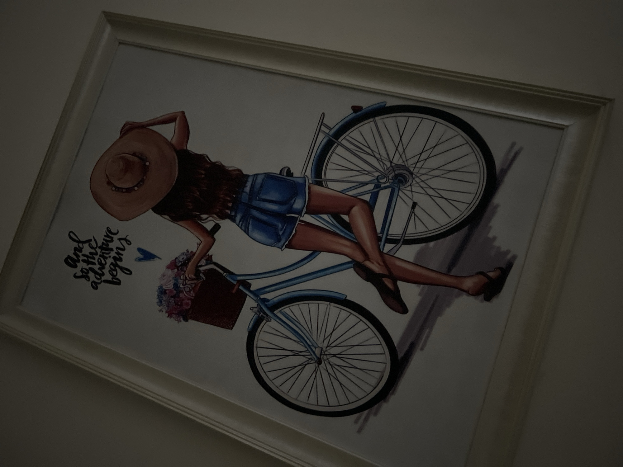 A low-light, angled photograph captures a framed print, likely mounted on a wall. The white or light-colored frame encloses an illustration of a stylized female figure in a relaxed, reclining pose atop a light blue bicycle. The woman has long, dark brown hair and wears a wide-brimmed straw hat with beaded details, denim shorts, and dark high-heeled sandals.
The bicycle features silver spokes, black tires, and a brown basket attached to the handlebars, overflowing with pink, purple, and white flowers. The background of the illustration is plain white, with subtle grey shading beneath the bicycle, possibly indicating a shadow or ground. To the left of the figure, decorative text reads: "And so the adventure begins," accompanied by a small blue heart.
The overall impression from the illustration suggests themes of leisure, travel, and new beginnings. The photograph's dim lighting and oblique angle slightly obscure some details of the print. The image itself does not contain any visual cues to specify its location in Gwarinpa, Nigeria.