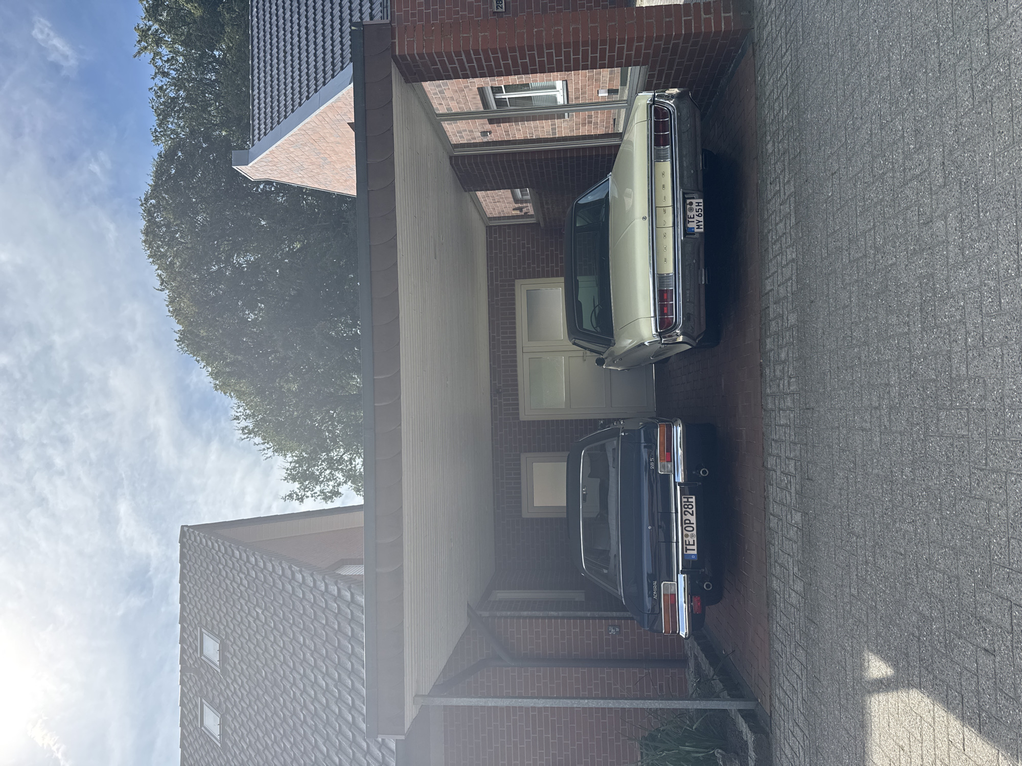 The image shows a carport with two classic cars parked underneath. The carport is attached to a brick house. The house has a tiled roof with two dormer windows. There is a large tree behind the house. The car on the left is dark blue and has the license plate "TE OP 28H". The car on the right is cream-colored and has the license plate "TE MY 65H". Both cars appear to be in good condition. The pavement in front of the carport is made of interlocking bricks. The sky is blue with some white clouds. The sun is shining. The location appears to be in Rheine, Germany, as suggested by the license plates.