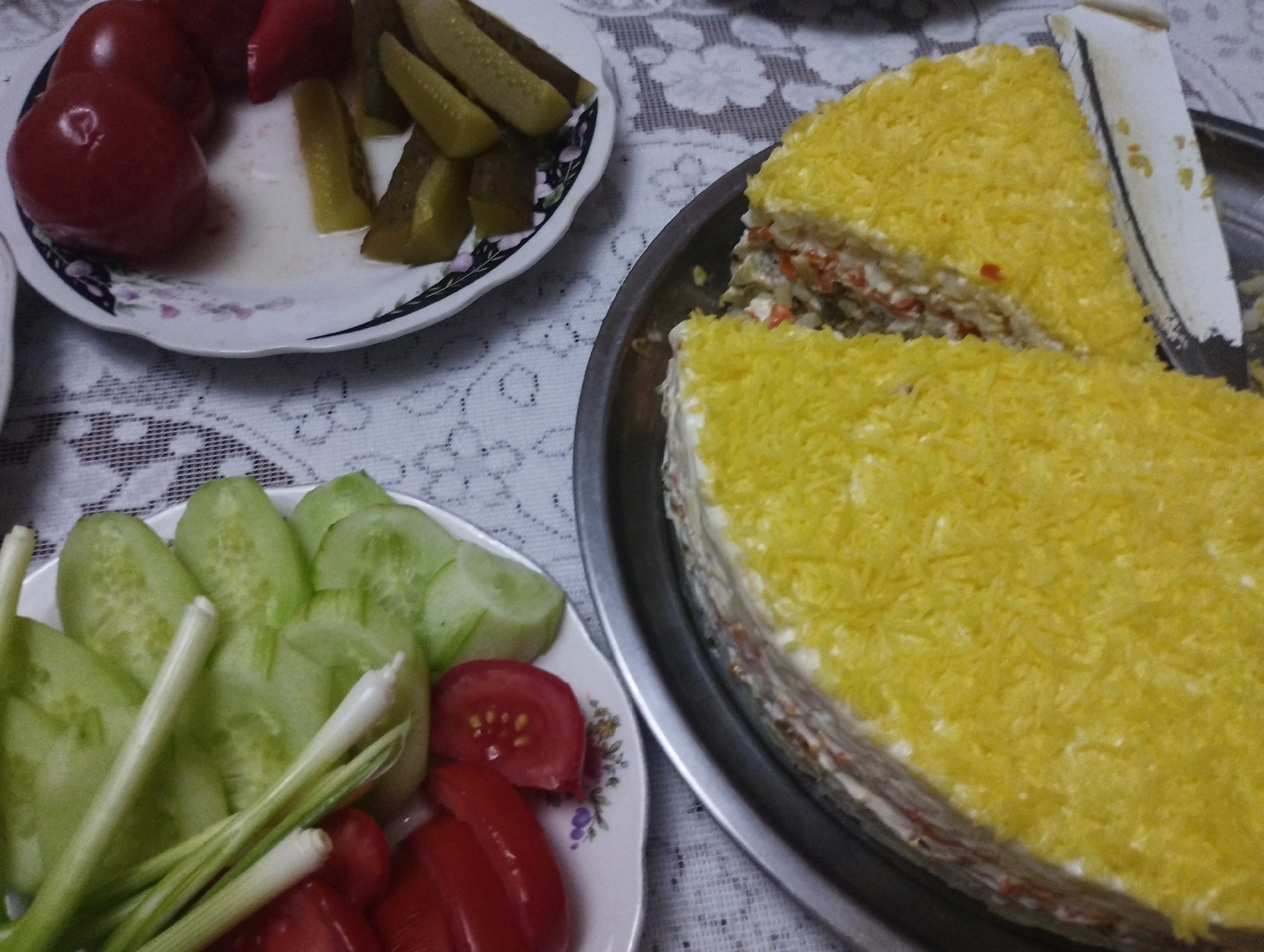 A close-up, overhead shot captures a portion of a meal laid out on a white, lace-patterned tablecloth. To the left, a white plate displays sliced tomatoes, pickles, and what appear to be green onions. Next to it, a serving dish holds sliced cucumbers and more chopped tomatoes. Dominating the right side of the frame is a sliced, multi-layered cake, topped with finely grated yellow material, likely cheese or egg yolk. A knife with a serrated edge is partially inserted into the cake, suggesting it has been cut. The lighting is soft, casting minimal shadows. The setting appears to be a domestic dining area, possibly in Bilajari, Azerbaijan, given the context provided.