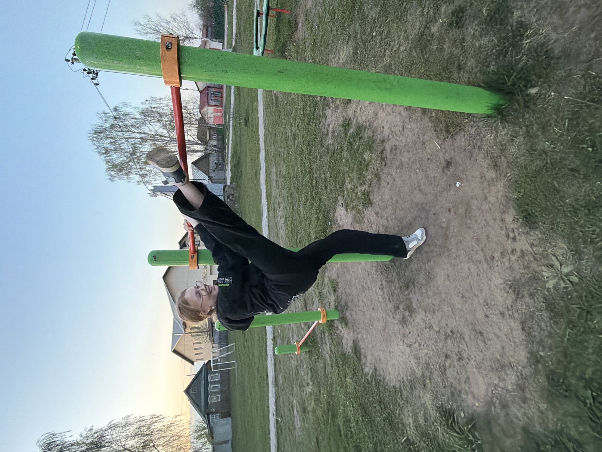The image shows a woman performing a gymnastic exercise on outdoor fitness equipment in what appears to be a park or recreational area. She is hanging upside down, supported by the equipment, with one leg extended straight up and the other bent. The woman is wearing dark clothing and glasses.
The fitness equipment is constructed of green bars with orange supports. The ground around the equipment is a mixture of dirt and grass. In the background, there are buildings, including houses, suggesting this location is within a residential area. Trees are visible in the background, contributing to the park-like environment. The lighting suggests it is late afternoon or early evening, indicated by the sky's color.
This location is specified as Chaikovsky, Russia.