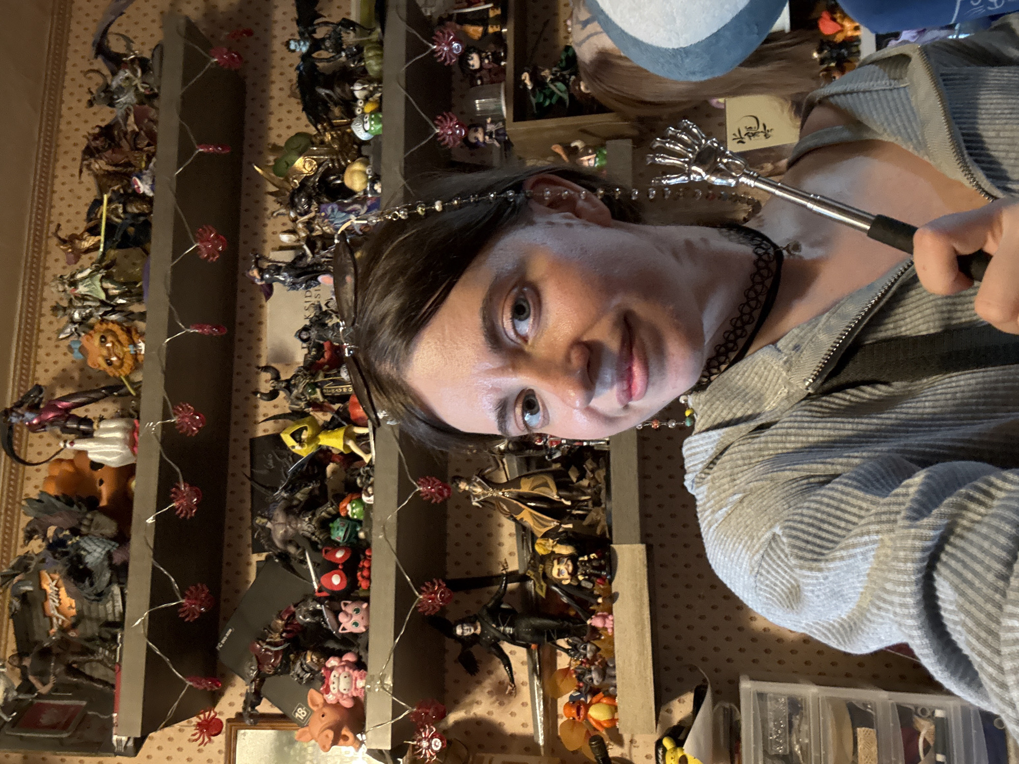 The image shows a person holding a silver, extendable rod with a hand-shaped end. The person has dark hair and is wearing a grey zippered jacket and a black choker. Behind the person is a wall-mounted shelf filled with numerous figurines. The background wall has a patterned wallpaper. There is a string of lights with red, spiky decorations intertwined with the shelves. The image does not have enough information to confirm if it was taken in the city of Moscow, Russia.