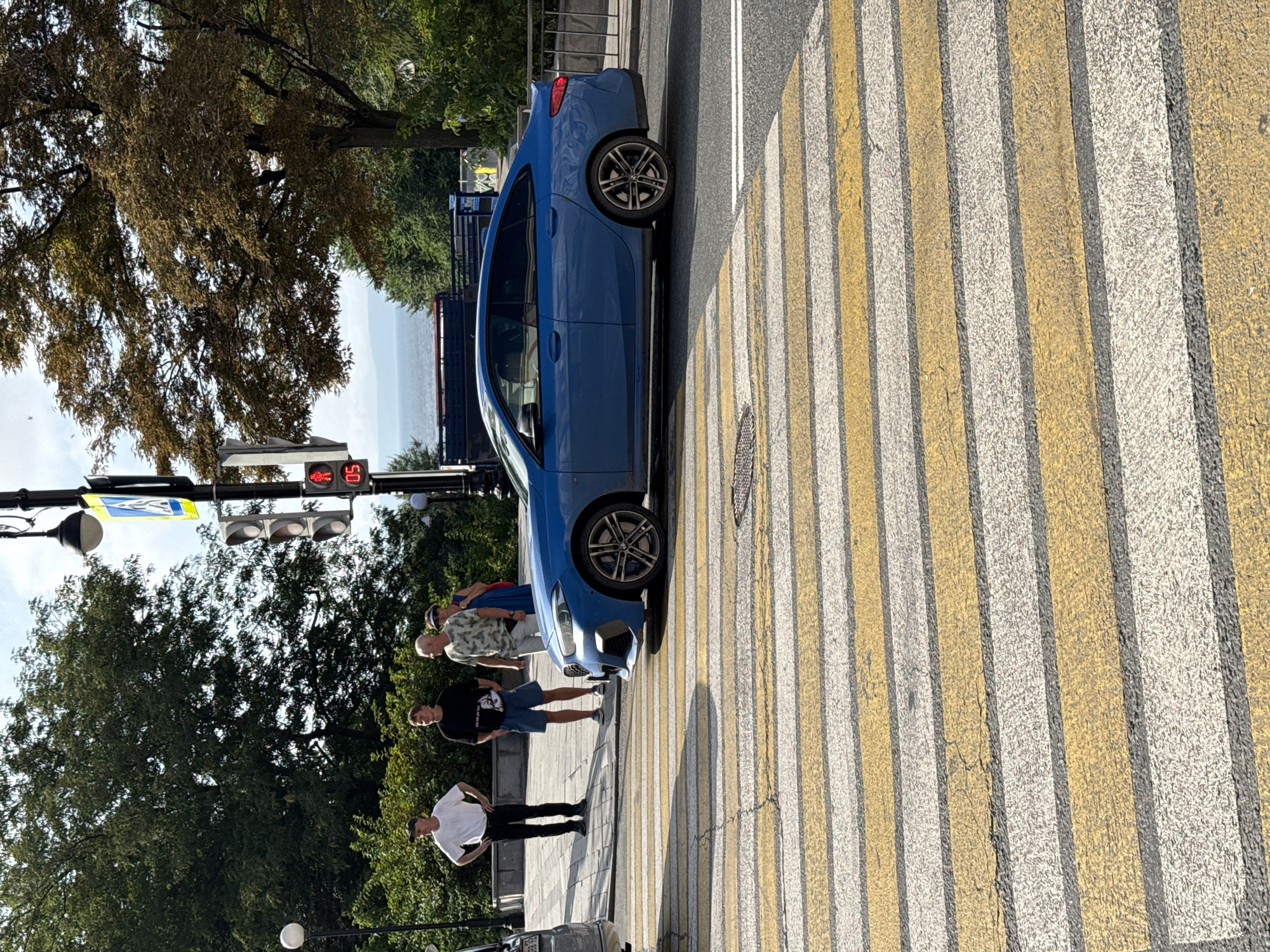 A blue BMW 2 Series Gran Coupe is stopped at a pedestrian crossing on a sunny day in Vladivostok, Russia. Four people, two men in casual attire, one man in a t-shirt and shorts, and a woman in a blue dress, stand on the sidewalk to the left of the car. The car's front left wheel is on the pedestrian crossing, which is marked with yellow and white stripes. Behind the car, a busy street with trees and buildings can be seen. In the background, a body of water and a distant shoreline are visible. A traffic light with a red pedestrian signal is also visible.