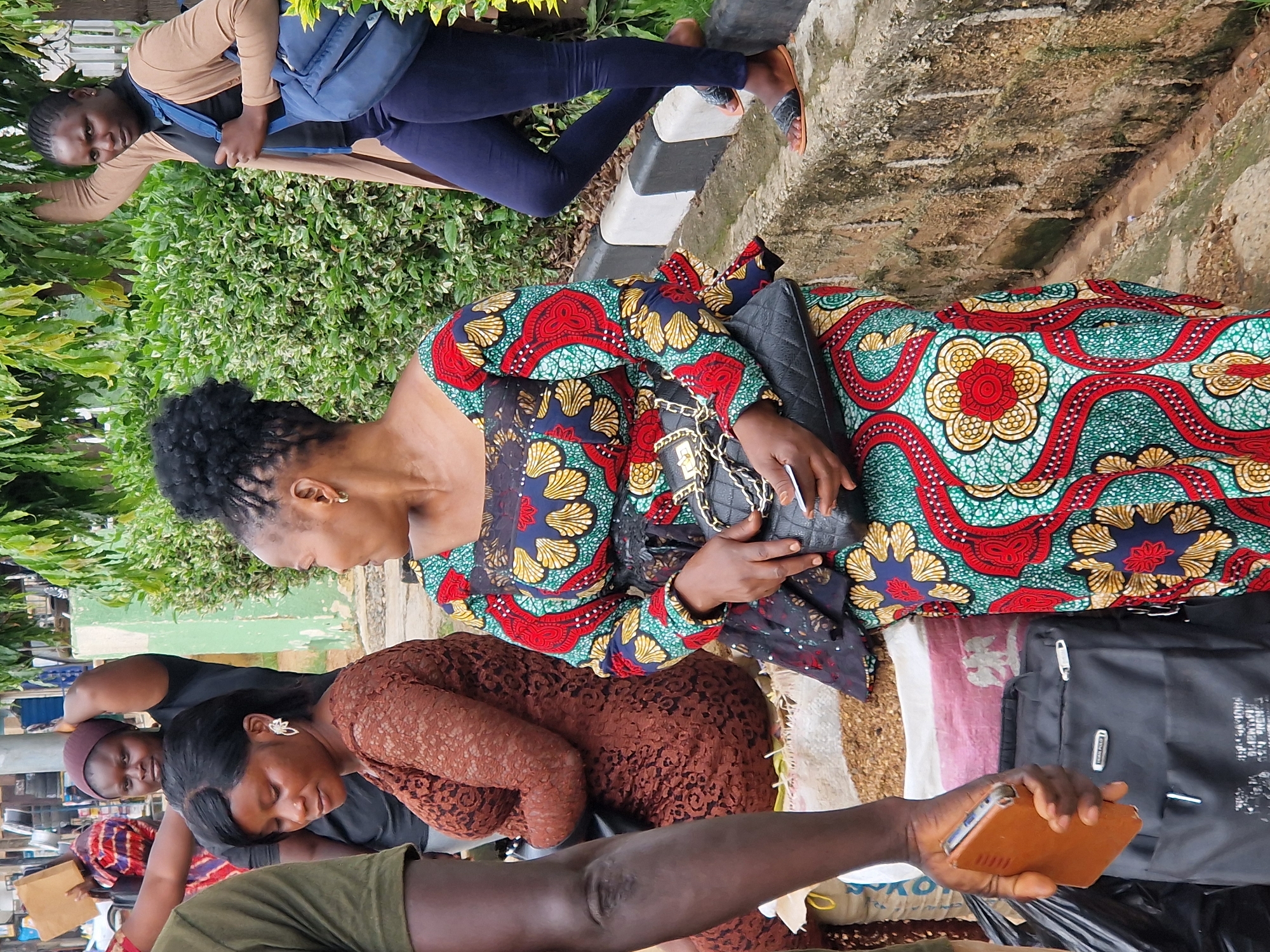 In Kabong, Nigeria, a group of people are gathered outdoors near a street lined with lush green bushes. A woman in the foreground, wearing a vibrant, patterned dress with red, yellow, and blue floral motifs, is seated. She is holding a black quilted handbag and a brown smartphone. To her left, another woman in a brown lace dress is also seated. Behind them, a woman in a black t-shirt and maroon headscarf is visible, and further back, another woman in a beige cardigan and dark blue leggings stands with a blue backpack. The ground is covered with bags of produce, and a black backpack lies near the foreground. A stone wall borders the area.