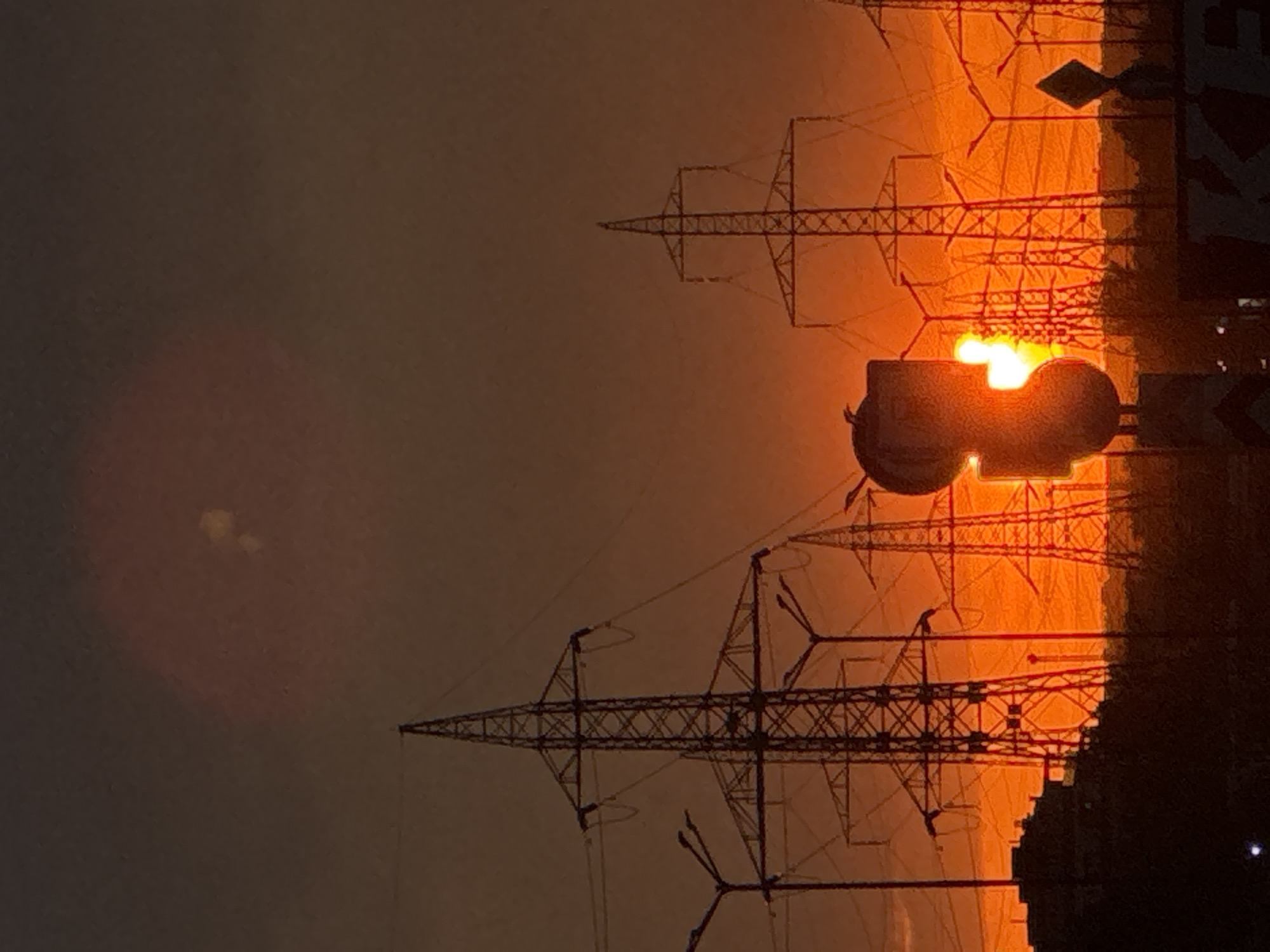 The image shows a landscape with power lines, with the sun setting behind them. The scene is likely taken from Sofia, Bulgaria. The power lines are tall, metal structures. The sun is setting, creating an orange and yellow glow in the sky. The city skyline is visible in the background.