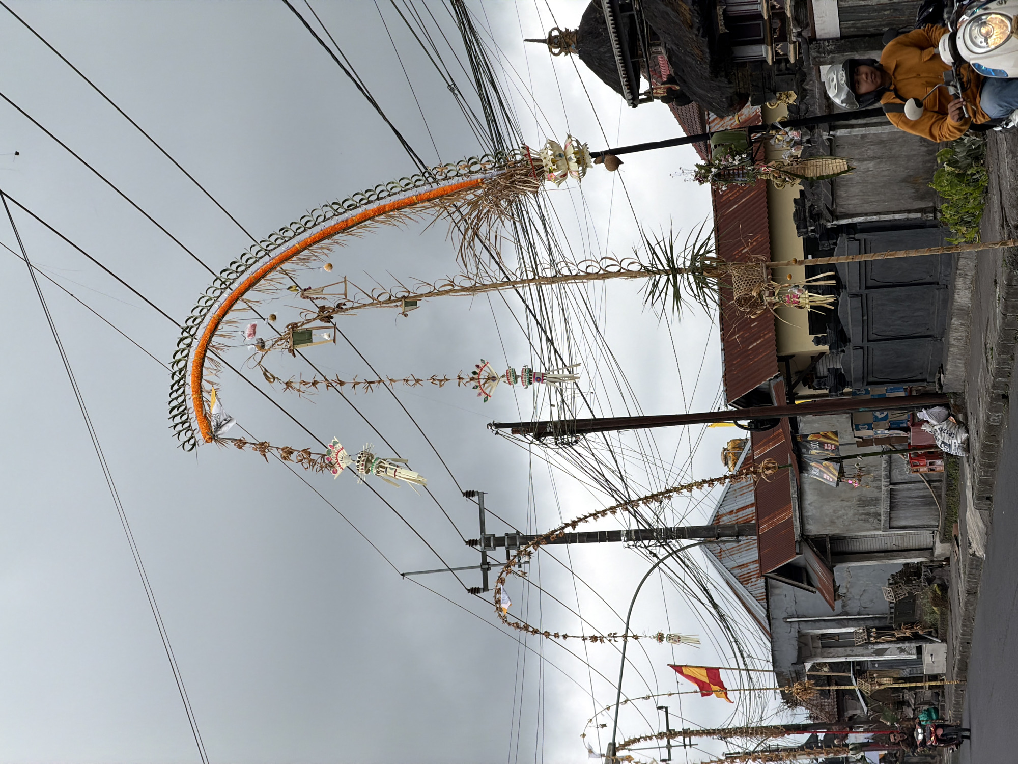 In Kintamani, Indonesia, an overcast sky hangs above a street scene adorned with festive decorations. A prominent, arching bamboo structure, decorated with orange flowers and white and green tassels, spans across the street, partially obscured by a dense network of power lines. Smaller, hanging decorations of a similar style are also visible, suspended from these lines. On the left side of the street, a road runs parallel to a row of buildings with corrugated iron roofs and weathered facades. A man wearing a yellow jacket and a helmet rides a white scooter towards the viewer, his face visible. To the right, a traditional Balinese building with a thatched roof and intricate carvings is partially visible. Various bags of what appear to be rice or cement are stacked near the roadside, alongside a dark fence. The overall impression is a blend of everyday urban life and cultural celebration.