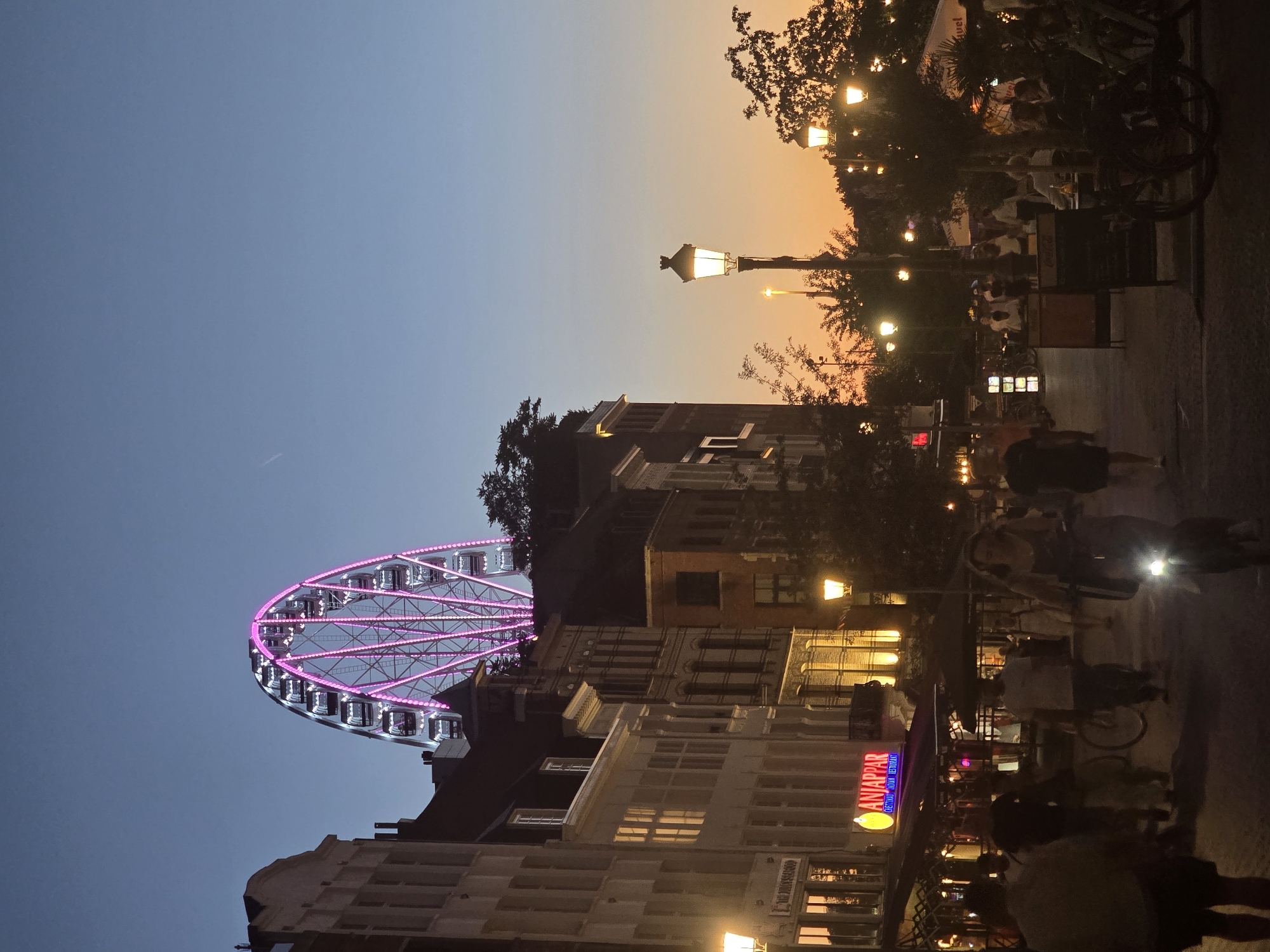 The image depicts a street scene in Antwerp, Belgium, at dusk. A prominent Ferris wheel with pink illumination is visible in the background, towering over buildings. The street is lined with establishments, including one with a sign reading "ANJAPPAR CHEETTINAD INDIAN RESTAURANT." Several people are walking or cycling along the street, some of whom are illuminated by streetlights and the lights from the buildings. The sky shows a gradient of blue and orange, indicative of the twilight hour. The overall atmosphere is that of a lively urban environment transitioning into the evening.