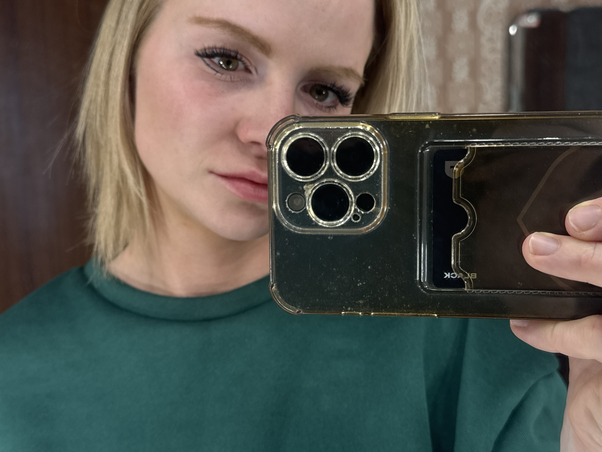 A selfie photograph depicts a fair-skinned woman with blonde hair, wearing a green long-sleeved shirt. She is holding a smartphone with a protective case in front of her face. The phone case obscures part of her face and body. The background appears to be indoors, with a blurred wall and a partial reflection of an object that looks like a mirror. The photograph does not depict any landmarks or other elements that could be used to determine a location in Saint Petersburg, Russia.