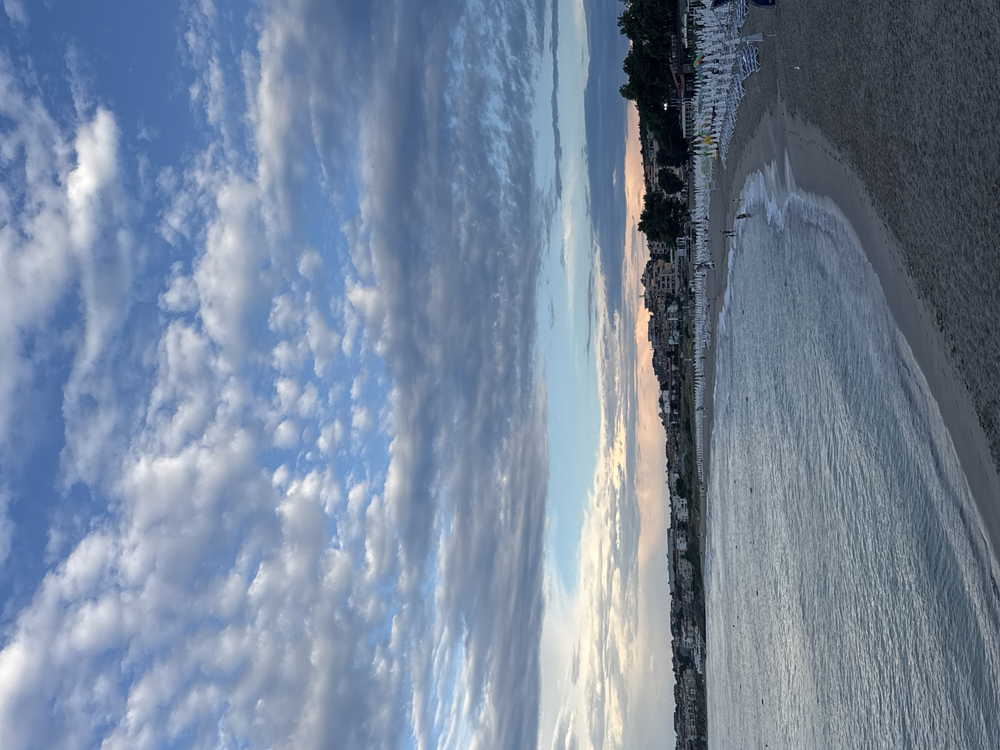 The image shows a view of the beach in Sozopol, Bulgaria. The sky is filled with a variety of clouds, ranging from dark, textured formations to lighter, wispy patterns against a bright blue background. In the distance, a coastline is visible with buildings and trees lining the shore. The beach itself is lined with numerous white lounge chairs and umbrellas, indicating it is a popular spot. The sea is calm with gentle waves rolling onto the sandy shore. A few people are present on the beach and in the water, engaged in casual activities. The overall scene depicts a serene coastal landscape during what appears to be late afternoon or early evening, with soft lighting.