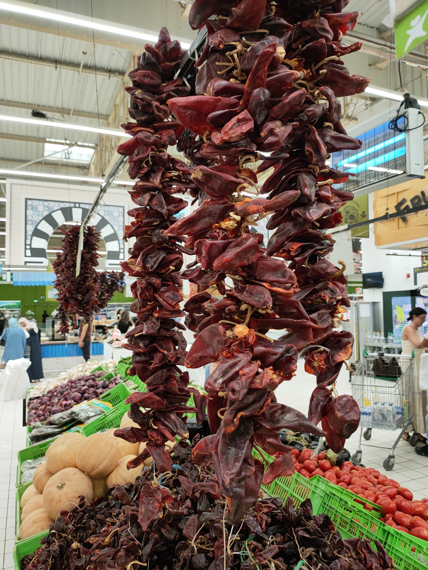 The image depicts a grocery store produce section with an abundance of dried chili peppers hanging from the ceiling. In the foreground, several strings of dark red, dried peppers are prominently displayed. Below them, green plastic crates are filled with various produce. One crate contains several large, light-colored pumpkins. Another crate holds a large quantity of red tomatoes. Further back, more green crates are visible, holding what appear to be red onions and other vegetables.
In the background, other shoppers are present. Two women, one in a blue hijab and a long blue garment, and another in a light-colored hijab and a dark dress, are standing near a produce display. Other individuals are seen browsing or pushing shopping carts. The store environment is characterized by bright overhead lighting, a tiled floor, and shelving units. A decorative archway with Islamic geometric patterns is visible on a wall in the mid-ground. The overall impression is a bustling supermarket produce aisle. The location is specified as Cebalat, Tunisia.