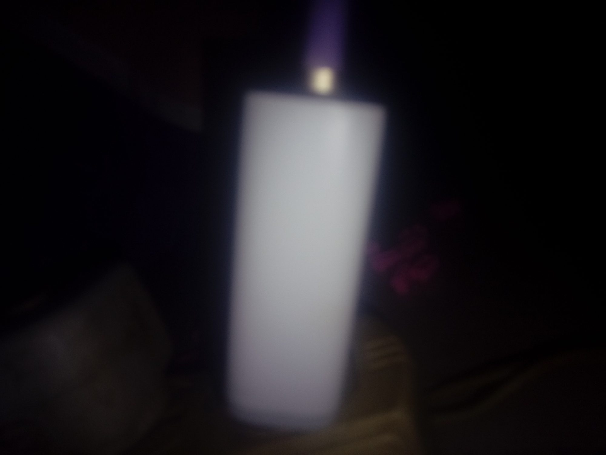 A blurry, low-lit photo shows a white, cylindrical object, possibly a bottle or container, with a purple top. The object is standing on a surface of unknown material. The background is dark and obscured, but vague shapes suggest surrounding objects. The location and specific details cannot be determined.