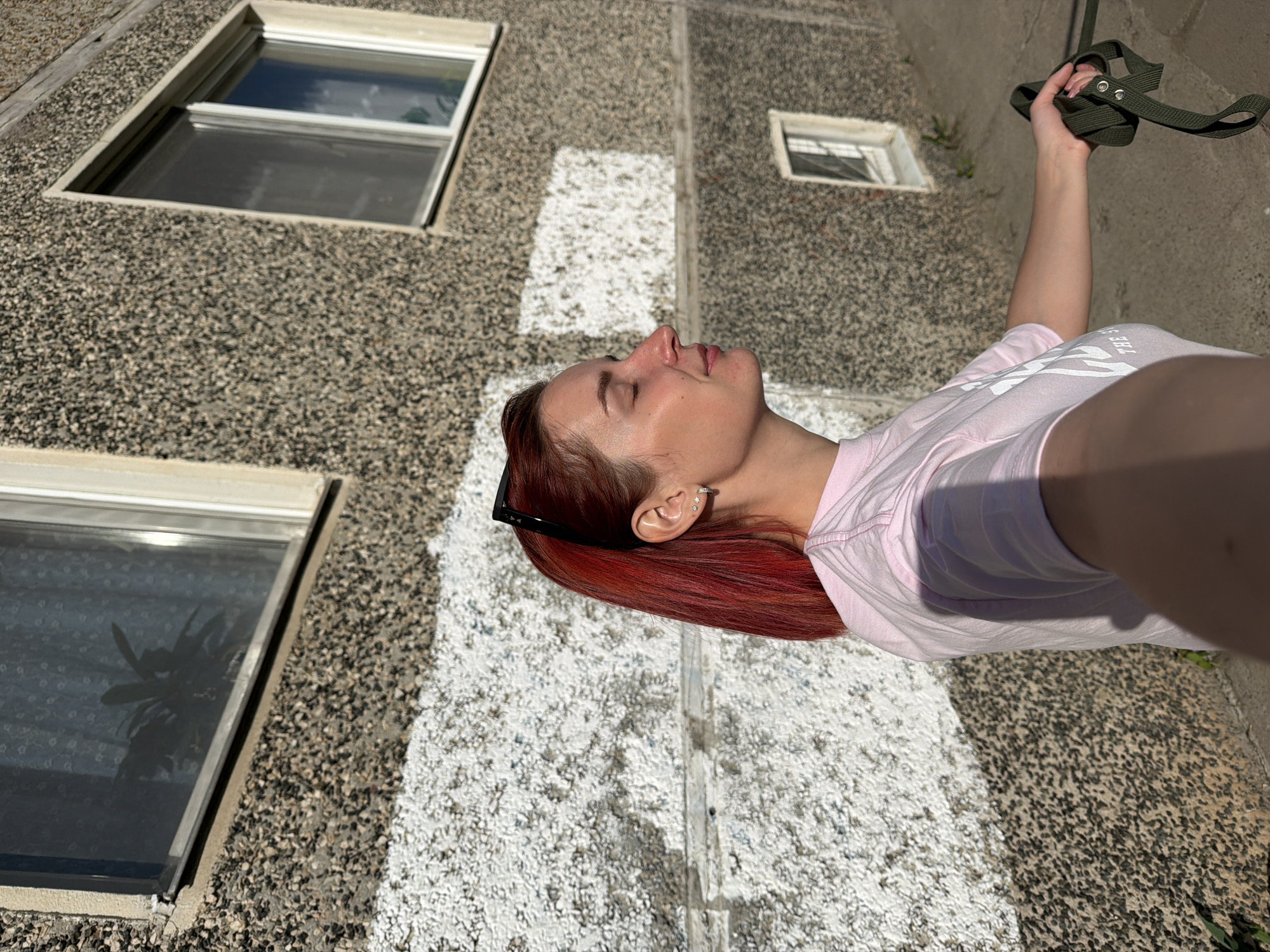 A medium shot depicts a woman standing outdoors in the city of Chaikovsky, Russia. She is holding a green strap with metal hardware in her raised right hand. The woman has red hair, sunglasses perched on her head, and a light pink t-shirt. Her face is turned upwards with her eyes closed, and she appears to be in sunlight. Behind her is a building with a pebble-dashed exterior and two visible windows. One window has a dark reflection of a plant inside. A vertical white painted stripe runs down the building's exterior. The ground beneath her feet appears to be concrete.