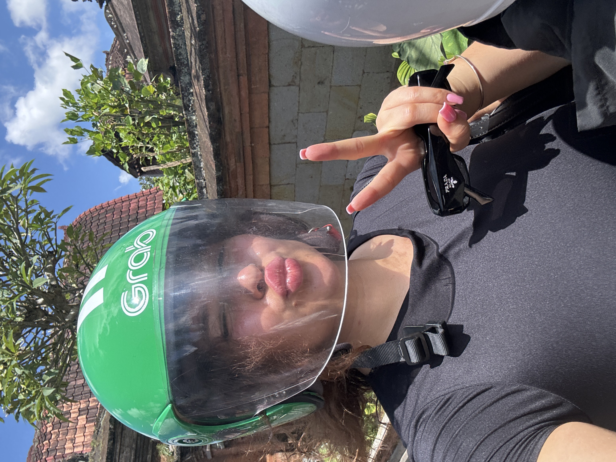 A person wearing a green Grab-branded helmet and black clothing is pictured making a peace sign while holding black sunglasses. The scene is outdoors in the Ubud District of Indonesia, with a view of a building with a tiled roof and greenery in the background. Another helmet, white in color, is visible in the upper portion of the frame. The individual is the main subject, and the background indicates a specific location.