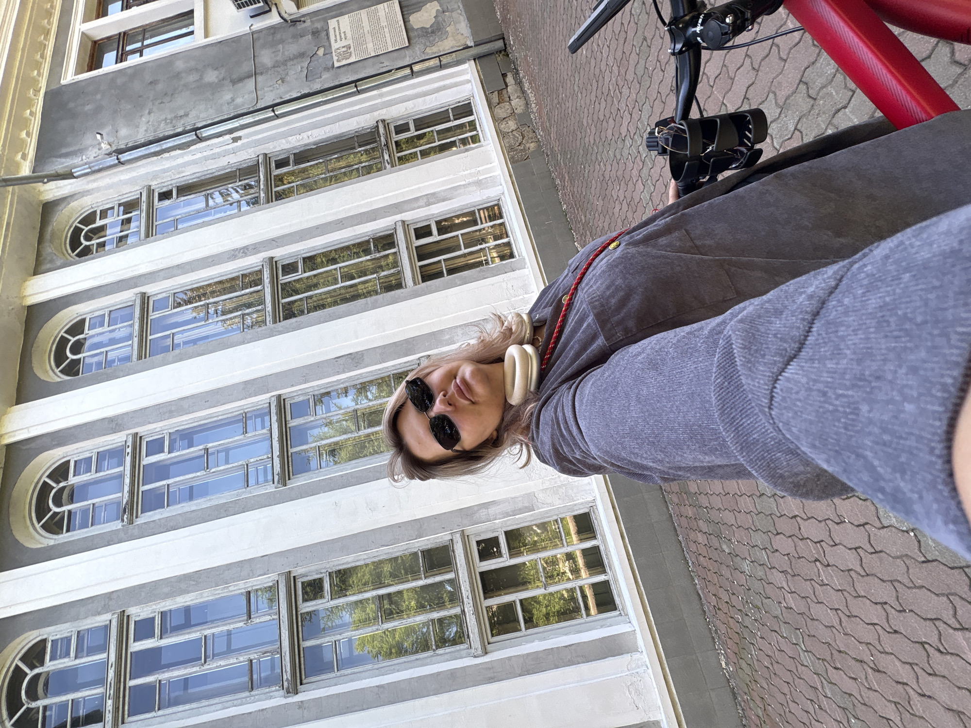 A woman is standing on a paved walkway in front of a building in Birobidzhan, Russia. The building appears to be old and has large, multi-paned windows. The woman is wearing sunglasses, a grey jacket, and a red cord around her neck. She is holding a camera, as if taking a selfie. A red bicycle is partially visible in the foreground. The walkway is made of interlocking red bricks. The overall lighting suggests it is daytime.
