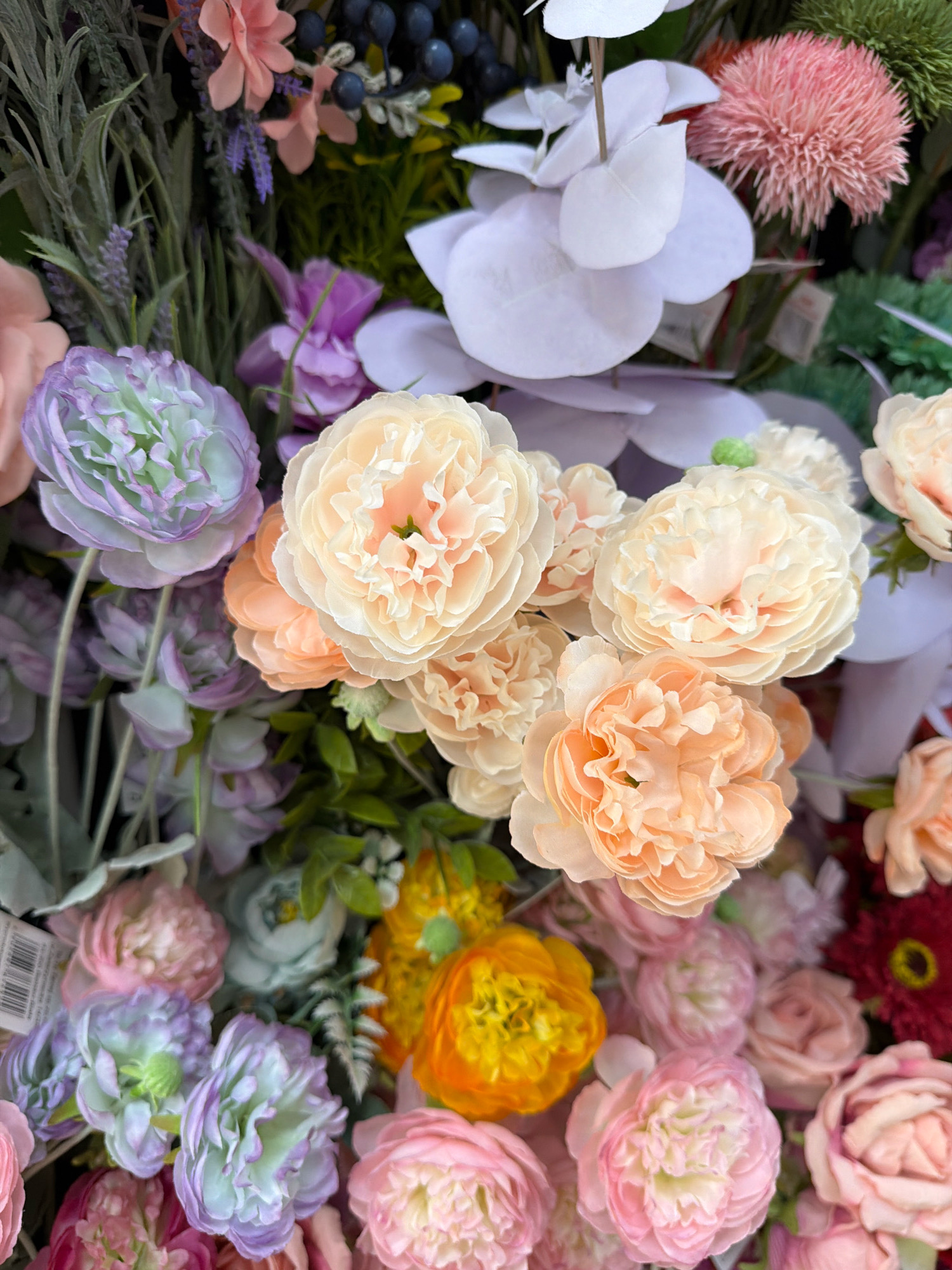 The image shows a close-up view of a vibrant assortment of artificial flowers. The flowers are in various shades of pink, peach, lavender, and yellow, with some greenery and dark berries interspersed. The arrangement appears to be a floral display or bouquet, with intricate petal details and layered textures. The lighting is bright and even, highlighting the colors and forms of the flowers. There are no people or discernible actions taking place. The context of Wrocław, Poland, is not visually evident in the provided image; it solely depicts a collection of artificial flowers.