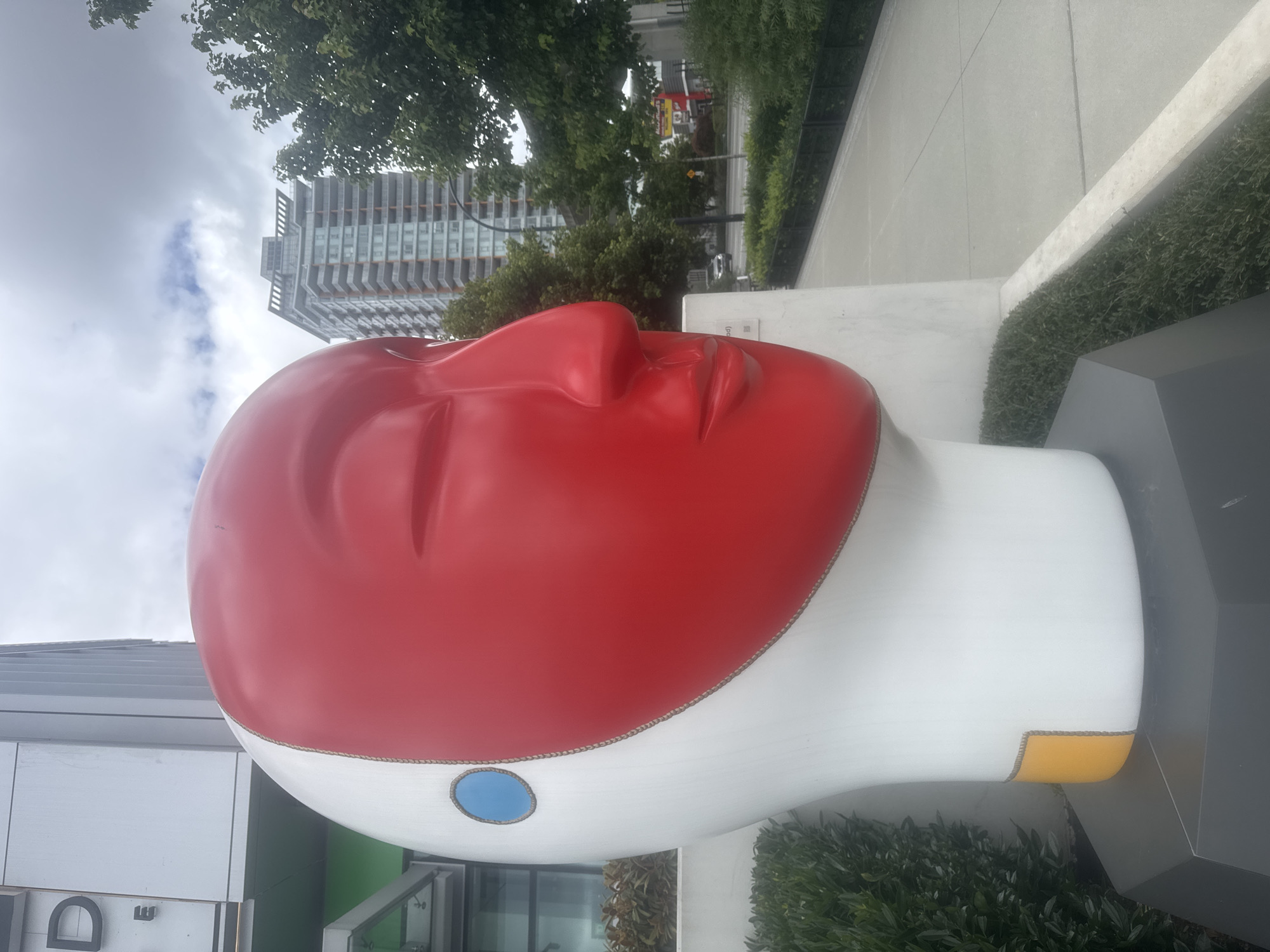 The image depicts a large, stylized sculpture of a human head situated outdoors in Burnaby, Canada. The sculpture is predominantly white, with a striking red face that covers the eye, nose, and mouth area. A small, circular blue detail is visible on the left side of the head, and a yellow rectangular detail is present at the base of the neck. The sculpture is placed on a grey, multi-faceted base.
Behind the sculpture, a modern building with multiple stories and balconies is visible. To the right, there are trees and a street with a few cars. The sky is overcast with clouds. The sculpture appears to be located in a public space, possibly near a building entrance or a walkway, indicated by the presence of manicured bushes and a paved pathway. The overall scene suggests an urban environment in Burnaby, Canada.