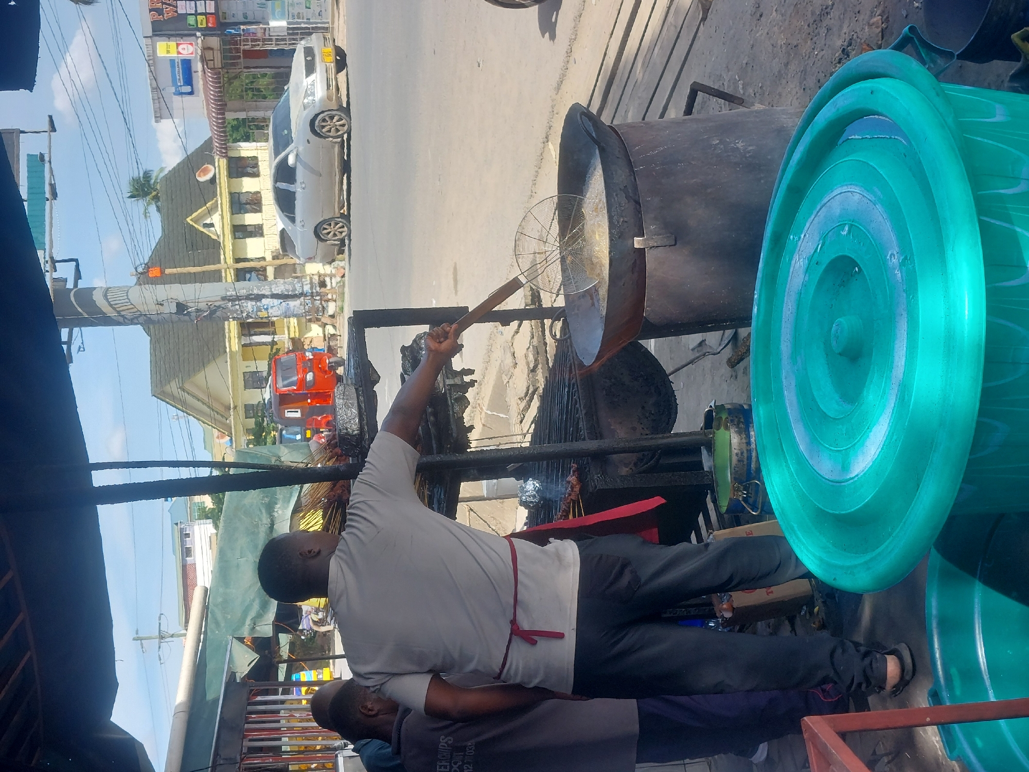 An outdoor food stall is located on a city street in Dar es-Salaam, Tanzania. A man in a gray shirt and dark pants, with a red apron around his waist, holds a metal strainer above a large, dark metal pot of hot oil. Fried food is presumably being cooked in the oil. Another man, wearing a darker shirt, stands beside the first man, near the front of the stall. A bright turquoise plastic tub is visible on the right side of the image. Other food items are cooking on a grill behind the man using the strainer. The street is visible in the background, with cars and buildings lining the street. An orange and white tricycle (bajaji) can be seen in the distance. Overhead power lines and poles are also visible in the image.