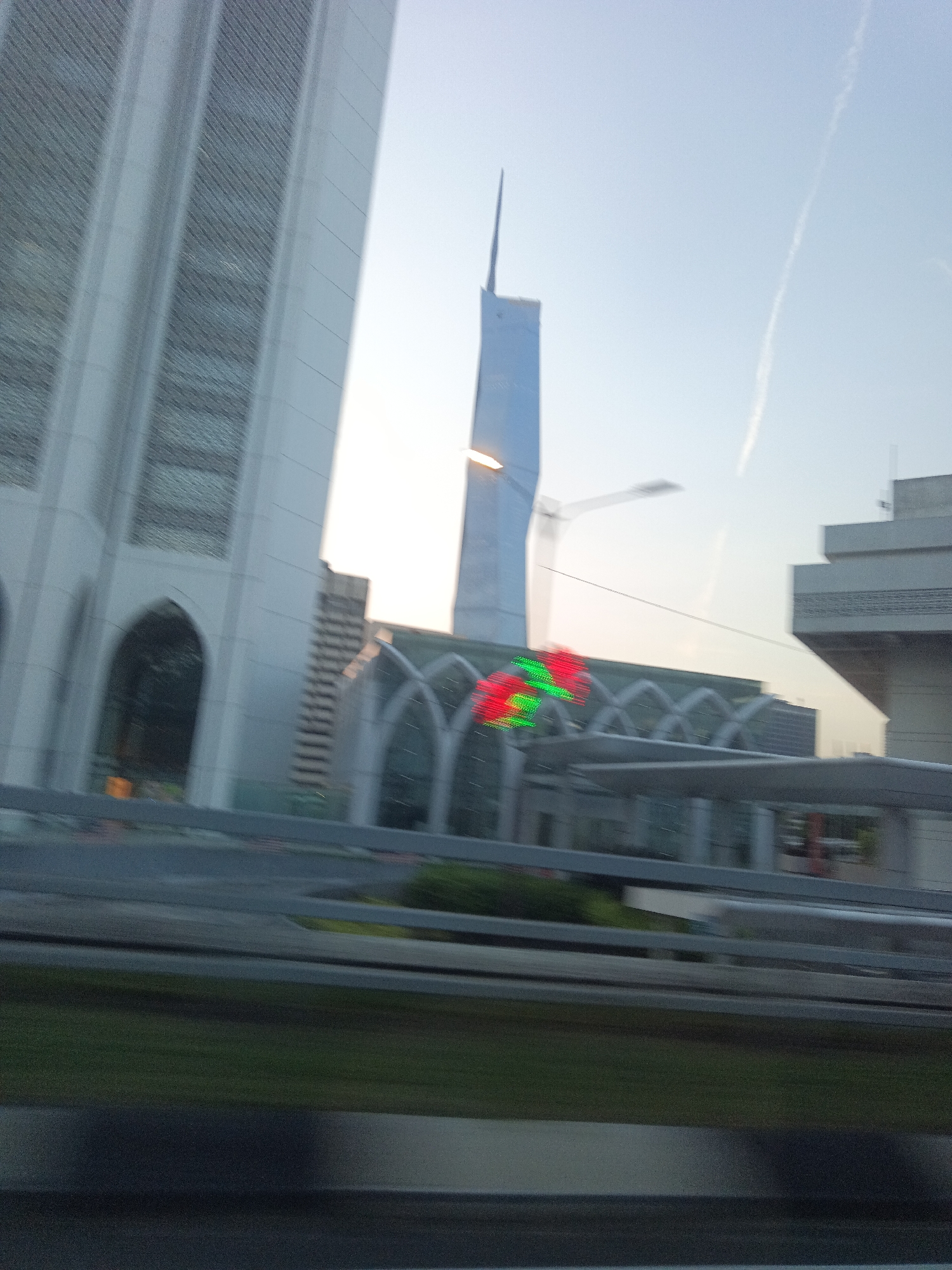 The image depicts a daytime scene of modern architecture in Kuala Lumpur, Malaysia, likely viewed from a moving vehicle. In the foreground, a blurred expanse of road and greenery suggests the vantage point. Dominating the midground and background are several distinct structures. On the left, a tall, angular building with vertical paneling, possibly a skyscraper, is partially visible. To its right, a distinctive, slender tower with a pointed top, reminiscent of a needle, pierces the sky. In the center, a low-lying building with a series of arched openings and a modern, curved roof structure is prominent. This structure appears to be a part of a larger complex, possibly a transportation hub or public facility. Red and green lights are visible on this central building, adding a splash of color. Further to the right, another multi-story building with a layered, stepped design is situated. The sky is a pale blue with a faint contrail from an aircraft. The overall impression is one of urban development and contemporary design within the city of Kuala Lumpur.