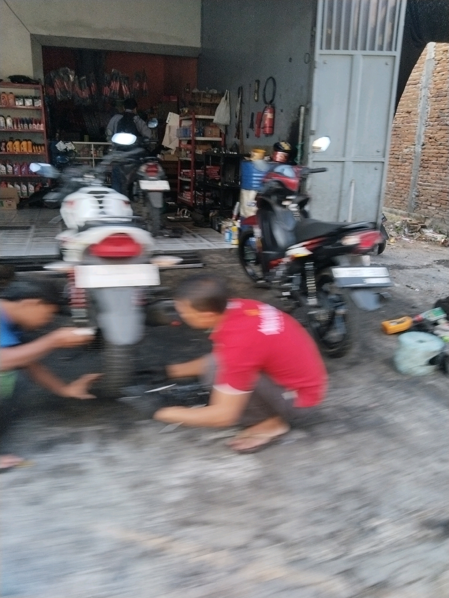 Two young males are working on a motorcycle in what appears to be a garage or workshop. The male on the left, wearing a blue shirt and green shorts, is crouched down and appears to be holding something near the motorcycle's rear tire. The male on the right, wearing a red t-shirt and gray shorts, is also crouched and seems to be attending to the same area of the motorcycle. The motorcycle itself is white with red accents and has a license plate. In the background, there are shelves stocked with various items, including bottles that could be lubricants or cleaning fluids. A second motorcycle, primarily black, is parked behind the white one. The ground is concrete and appears wet in places, suggesting it might be a car wash or a maintenance area. The overall environment is cluttered but functional.