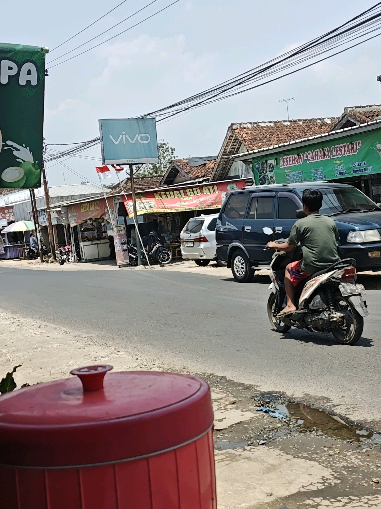 The image depicts a street scene in Kemuning, Indonesia, on a sunny day. In the foreground, a large red cylindrical object with a domed lid is partially visible. The street is paved, with a patch of rough, wet asphalt and debris to the right. A person is riding a white and red motorcycle away from the camera, wearing a green shirt and red and white checkered shorts. Behind the motorcycle, a dark blue minivan is parked, and further back, a white car is visible.
On the left side of the street, a green banner with white text "PA" is displayed. Next to it is a blue sign with "vivo" in white letters. Further down the street, several small shops and stalls line the road. One stall has a red and white banner that reads "KEDAI BU. AM". Another stall has a green banner with text that appears to be a menu. Indonesian flags are visible on poles near the "vivo" sign. Several motorcycles are parked in front of the shops. In the background, there are buildings with tiled roofs, and power lines crisscross the sky.
