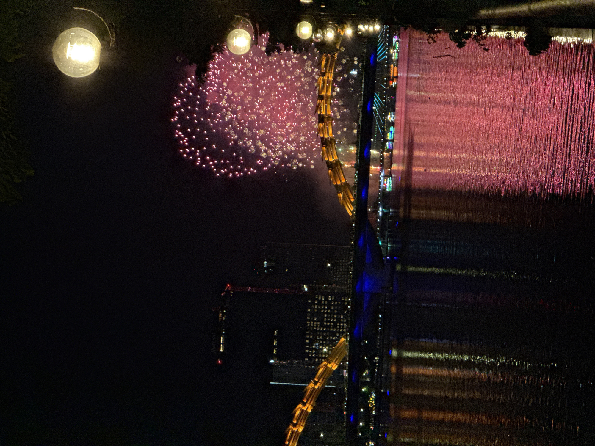 The image depicts a nighttime scene in Da Nang, Vietnam, likely during a fireworks display. The overall composition is oriented sideways, with the top of the image appearing at the bottom.
The foreground features a bright, circular light fixture, possibly a streetlamp or part of decorative lighting, casting a warm glow against the dark sky. To the right of the light, a cluster of pink and purple fireworks explodes, with numerous small, bright dots representing the individual pyrotechnic bursts.
Beyond the fireworks, a cityscape is visible, dominated by tall buildings illuminated with various colored lights, predominantly blue and white. A prominent, bright yellow structure, possibly a bridge or a large piece of architecture, curves diagonally across the scene, also lit up. In the background, a body of water reflects the colorful lights of the city and the fireworks, creating a shimmering, rippled effect. The reflection is a dominant feature, taking up a significant portion of the right side of the image. The water's surface appears textured, suggesting movement or ripples.
Further to the left and partially obscured, a construction crane is visible against the dark sky, with a red light at its tip. Some foliage can be seen in the upper left and upper center of the image, indicating a natural element juxtaposed with the urban environment.
The time of day is clearly night, characterized by the dark sky and artificial lighting. There are no visible people. The atmosphere suggests a festive or celebratory event due to the presence of fireworks and city illuminations. The reflection of the lights on the water adds a sense of depth and grandeur to the scene. No discernible text is visible in the image, apart from potentially a faint "Marriott" on a building in the lower left, but it is very blurry and unconfirmed.
