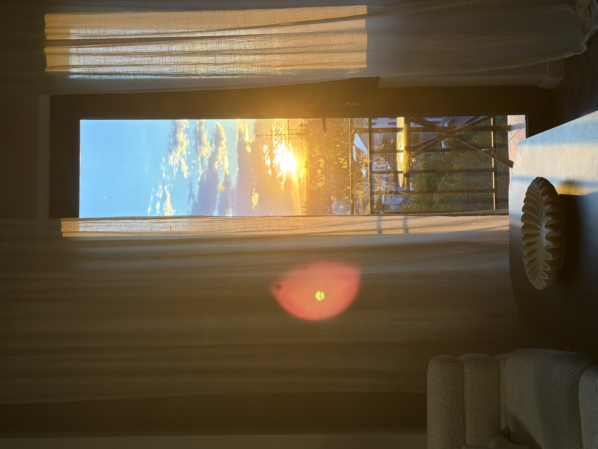 A vertical, eye-level, indoor shot depicts a sunset viewed through a window in Yerevan, Armenia. The scene is brightly lit with a golden hue from the setting sun, visible through the window. The window is partially covered by sheer, off-white curtains. Outside, the landscape includes a city view with buildings, trees, and an electrical transmission tower silhouetted against the vibrant sky.
A table with a decorative, off-white object is visible to the right. In the lower right, a textured, off-white chair is partially visible. A red glare is present in the lower-center of the frame.