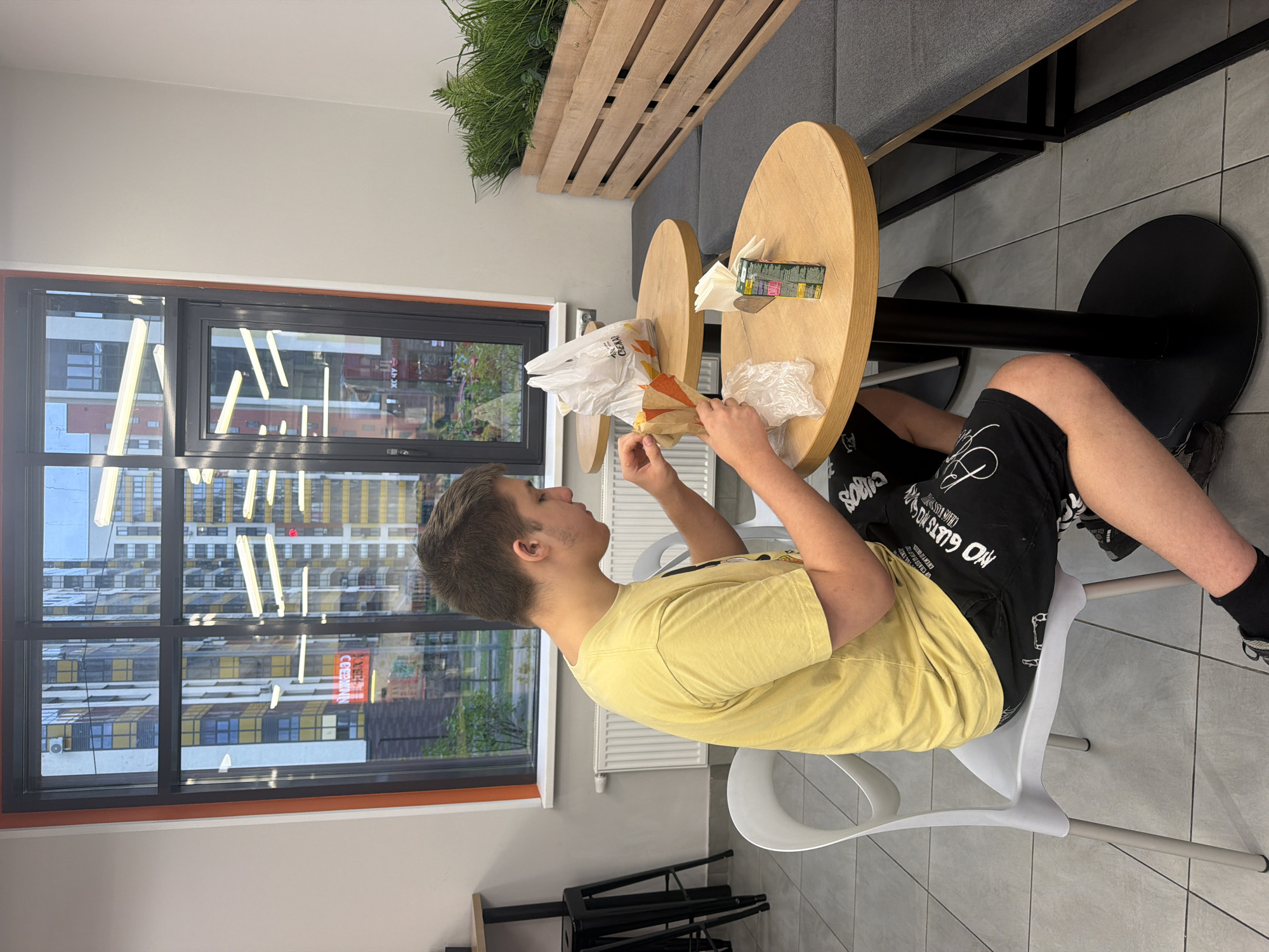 The image shows a young person sitting at a round, light-wood table in what appears to be a cafe or fast-food establishment. The person is wearing a yellow t-shirt and black shorts with white lettering. They are in the process of unwrapping a food item from a paper bag. On the table, there is also a white plastic bag, a small carton of juice, and a stack of napkins. In the background, there is a large window showing a cityscape with modern high-rise buildings. The reflection in the window includes what appears to be a neon sign with Russian text. To the right of the table, there is a long, cushioned bench seating area and a wooden planter box containing greenery. The floor is tiled. The overall scene suggests a casual dining environment within an urban setting.