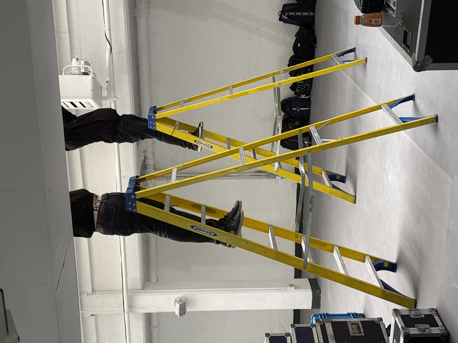 An indoor scene in what appears to be an industrial or event preparation space in London, United Kingdom, features two individuals standing on separate yellow Werner brand A-frame ladders, performing work on an overhead light grey horizontal structure.
The individual on the left ladder wears dark jeans, black sneakers, and a dark top, with a visible white waistband displaying "BOXY" branding above the trousers. The individual on the right ladder also wears dark trousers, black sneakers, and a dark top. Both individuals' upper torsos and heads are obscured by the overhead structure, indicating they are actively engaged in tasks above their visible height.
Above the right individual, a white rectangular HVAC unit or industrial fixture is mounted to the overhead structure. The surrounding environment consists of light grey concrete flooring and light grey/white walls with horizontal white pipes or conduits. A wall-mounted light fixture is visible on the left side. To the left, several black flight cases, some with blue accents, are stacked. In the mid-ground background, a collection of dark backpacks and bags are neatly arranged against the wall. On the far right, a section of a black flight case or table holds a brown plastic bottle with a label and a separate black cap.