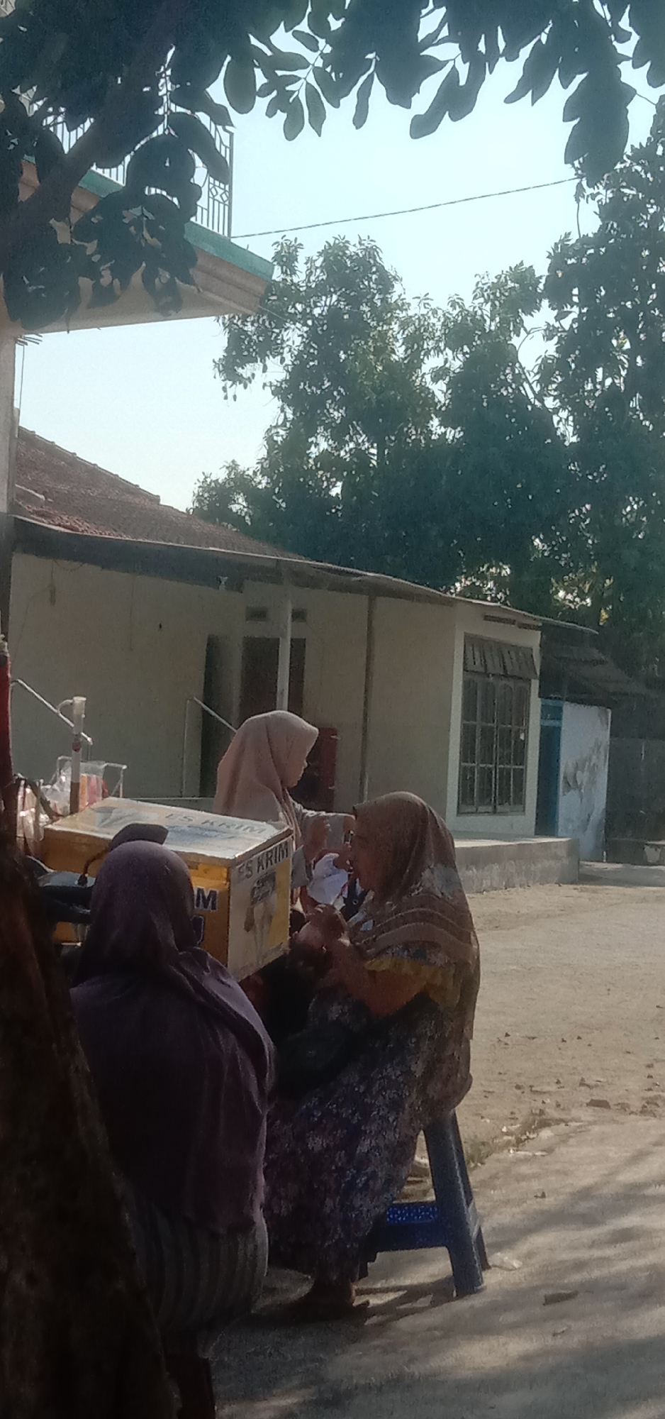 In a street scene in Sukorejo, Indonesia, three individuals are gathered around a portable ice cream cart. Two women, wearing hijabs, are the focus of the image. One woman, in a light pink hijab, appears to be serving or interacting with the other, who is in a patterned brown and yellow hijab and is seated. A third person, also wearing a hijab in a darker shade, is seated to the left and facing away from the viewer. The ice cream cart has "ES KRIM" visible on its side. In the background, a light-colored building with a tiled roof is visible, along with trees and a bright, possibly hazy, sky. The ground is paved with concrete or asphalt.