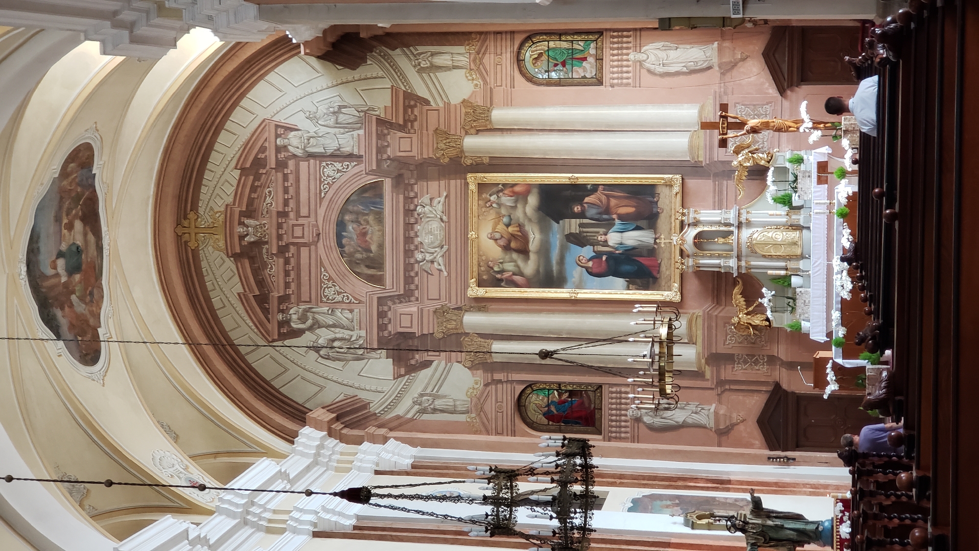 The image depicts the interior of a church in Prešov, Slovakia. The scene is a wide shot, capturing the ornate ceiling, altar, and pews. The architecture is grand, with a domed ceiling adorned with frescoes and detailed moldings. The walls are painted a pale pink, and the columns are white with gold detailing.
At the front of the church is a large altar, elaborately decorated with gold elements and white fabric. Above the altar hangs a crucifix with a golden figure of Christ and a golden angel at its base. To the left of the altar is a large framed painting depicting religious figures, and to the right, there are stained-glass windows. The pews are made of dark wood, and several people are seated in them, facing the altar. One person in the foreground is wearing a purple shirt and another in the background is wearing a white shirt. The lighting in the church appears to be a mix of natural light coming from the windows and artificial light from chandeliers. The overall mood is one of reverence and quiet contemplation.