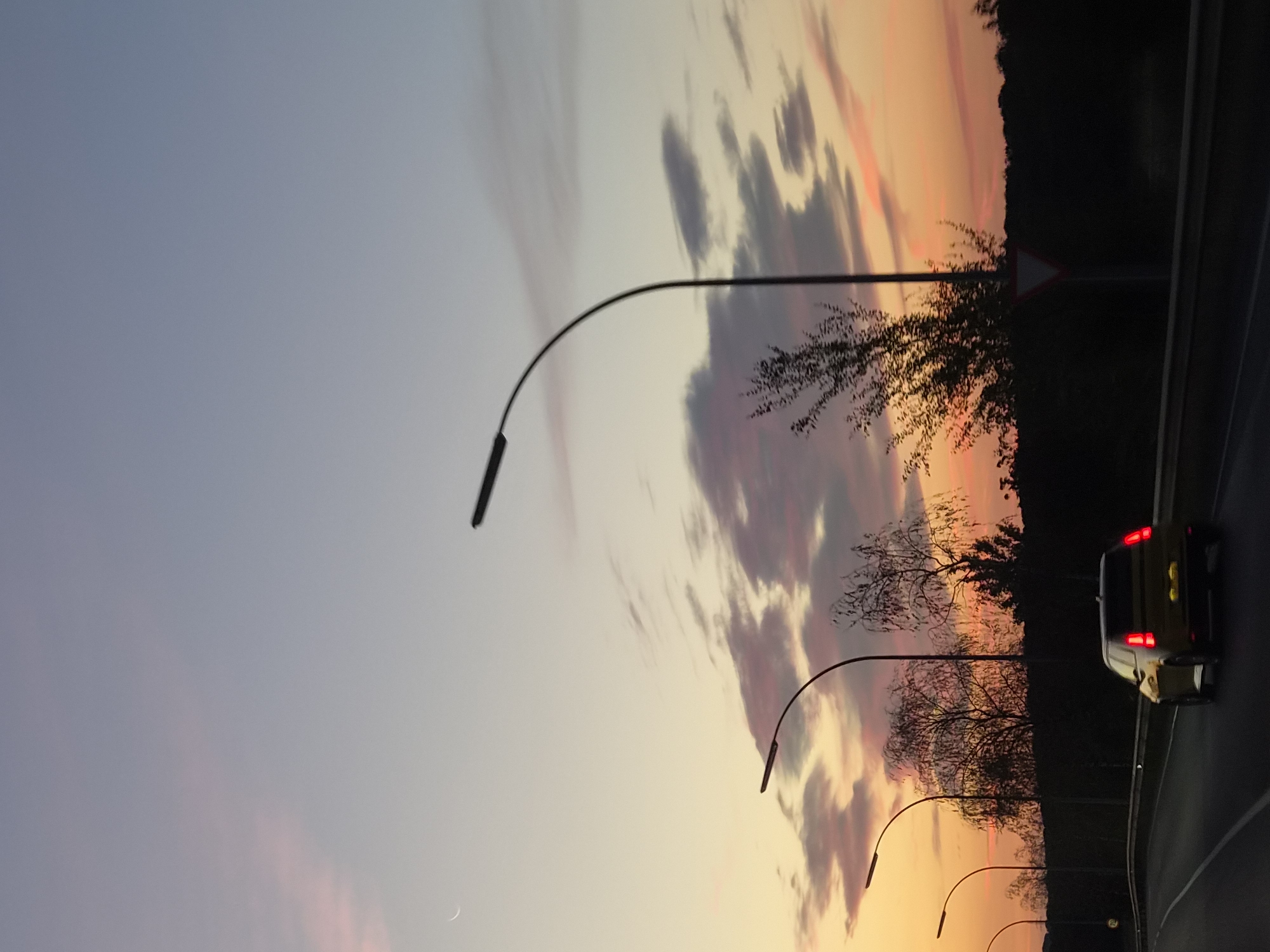 This is a low-angle, vertical, full shot of a road in Leudelange, Luxembourg, captured during a sunset. The sky displays a gradient of colors from a light blue at the top to hues of pink, orange, and yellow near the horizon. Wispy clouds are scattered across the sky, some tinged with pink and orange from the setting sun. A crescent moon is visible in the upper left quadrant of the sky.
In the foreground, a yellow car is driving away from the viewer on a multi-lane road. The car's brake lights are illuminated. The road is lined with a metal guardrail and streetlights with curved arms are positioned along the side of the road at intervals. In the background, a dense line of dark trees forms the horizon. A triangular road sign, possibly a yield sign, is visible to the right of the road, partially obscured by a bare-branched tree. The overall scene depicts a journey along a road at dusk.