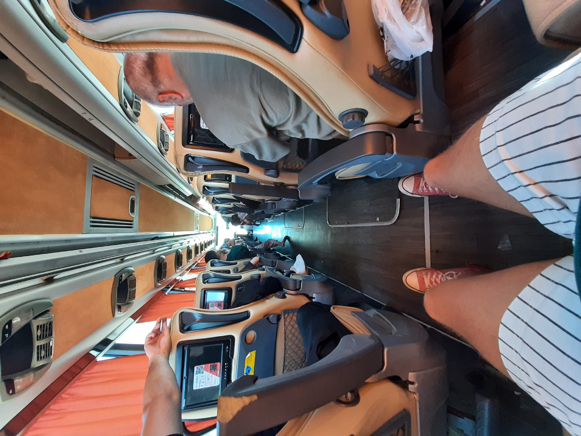 The image shows the interior of a bus, likely a sleeper bus, with passengers resting in reclined seats. The seats are upholstered in light brown leather and are arranged in rows on either side of a central aisle. Each seat appears to have individual entertainment screens.
In the foreground, a person's legs are visible, wearing striped shorts and red sneakers. Another person, a man with a bald head, is seen sitting in one of the seats, looking downwards. Further down the aisle, several other passengers are visible, some sleeping or resting in their seats. The bus appears to be dimly lit, possibly indicating nighttime or an enclosed environment. The flooring is dark wood. Orange curtains are visible along the sides of the bus.
Visible text includes a QR code and some text on a screen within one of the seats, but it is not clearly legible. The overall scene suggests a journey or a stopover on a long-distance bus trip. The presence of multiple passengers resting indicates the typical use of such a bus for overnight travel. The location is identified as Meram, Turkey.