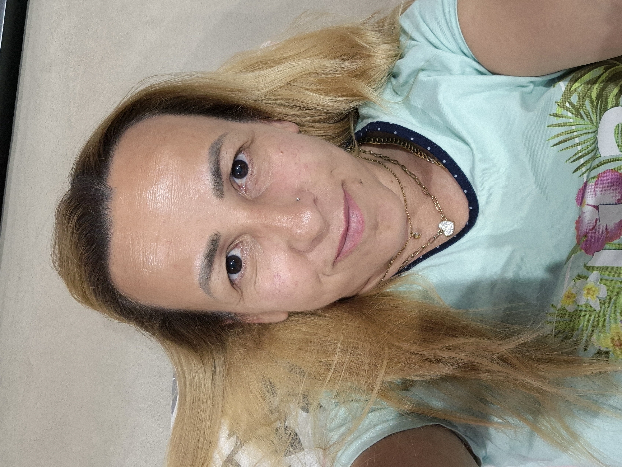 A selfie taken from a high angle shows a person with blonde hair lying down. The person is looking at the camera with a smile, wearing a light blue t-shirt with a floral pattern and a dark blue trim around the neckline. The person has a small nose ring and is wearing two gold necklaces, one with a heart-shaped pendant. The background is a neutral-colored, textured surface, possibly a wall or bedding.
