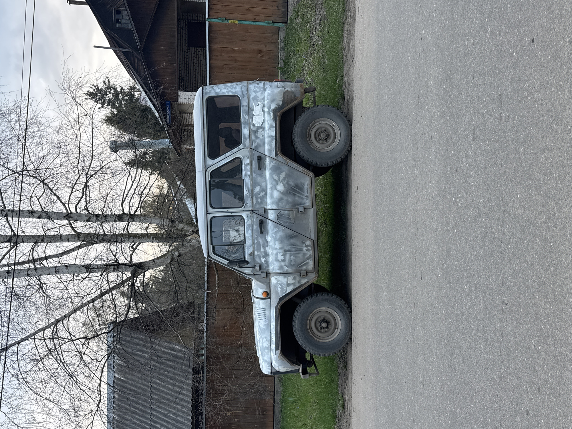 The image depicts a grey UAZ-469 parked on an asphalt road, likely in Tomilino, Russia. The vehicle's paint is partially removed, showing signs of rust and wear. It has large, off-road tires and a boxy, utilitarian design. Behind the vehicle is a wooden building with some peeling paint. To the left are bare birch trees and a building with a grey corrugated metal roof. The road appears to be a local street within the town. The lighting suggests it is daytime, likely early spring due to the leafless trees. A street sign in Cyrillic is visible on the wooden building.