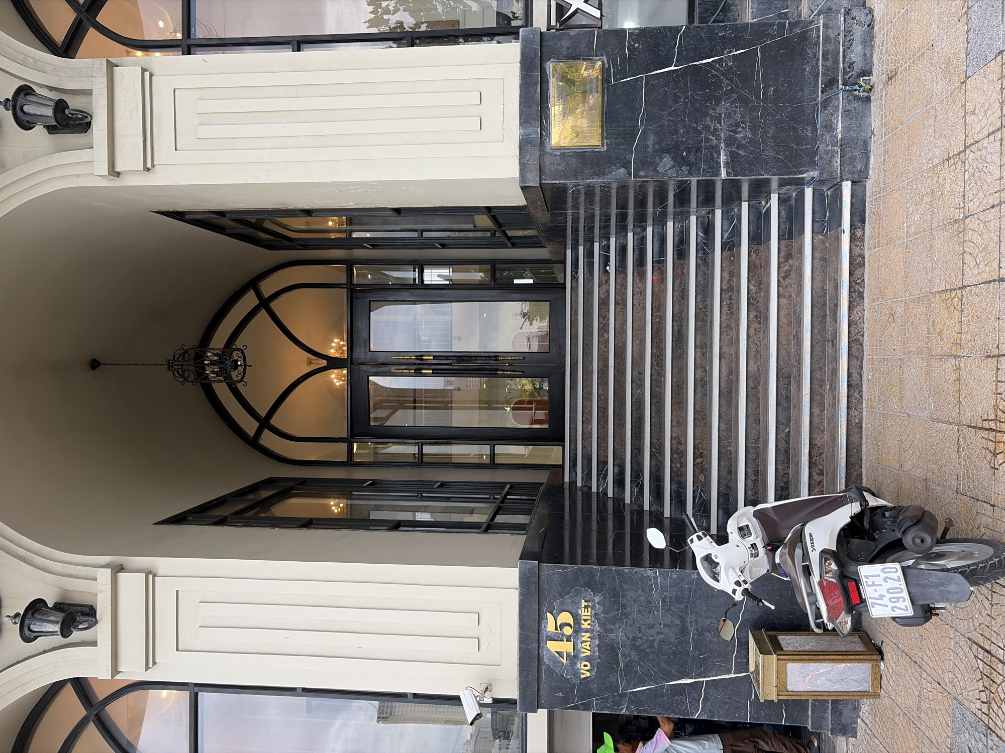 The image shows the entrance to a building located at 45 Vo Van Kiet Street in Da Nang, Vietnam. The entrance features a grand staircase made of black marble with white trim. Above the staircase, a set of double glass doors with ornate handles leads into the building. The building's facade is painted a cream color and is decorated with classical architectural elements, including large arched windows and decorative lamps.
In front of the staircase, a white scooter is parked. A person wearing a green hat and a light-colored shirt is partially visible on the left side of the image, standing near a golden decorative box. A security camera is mounted on the wall to the left of the entrance. A golden plaque is affixed to the black marble wall to the right of the entrance. The ground in front of the entrance is paved with patterned tiles. The sky appears overcast.