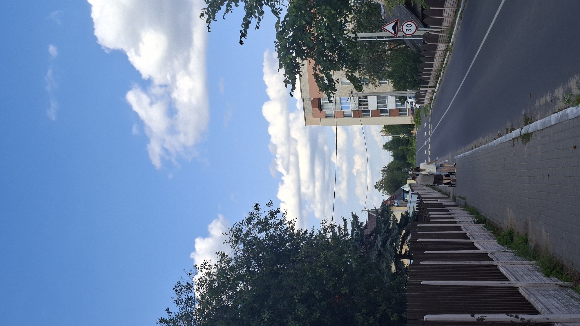 In Myadzyel, Belarus, a paved sidewalk runs alongside a dark asphalt road, bordered by a brown wooden fence with white concrete posts. Two individuals, a man and a woman, walk on the sidewalk pushing a stroller. They are dressed casually; the man wears a gray t-shirt and shorts, while the woman wears a white t-shirt and denim shorts. Behind them, a multi-story brick building stands on the right side of the road, with trees and other smaller buildings in the background. A traffic sign indicating a speed limit of 30 is visible near the building. The sky above is bright blue with scattered white clouds.